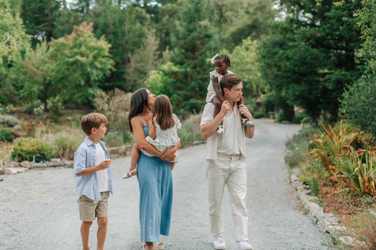 A happy family of five walks along a scenic garden path surrounded by lush green trees on a sunny day.