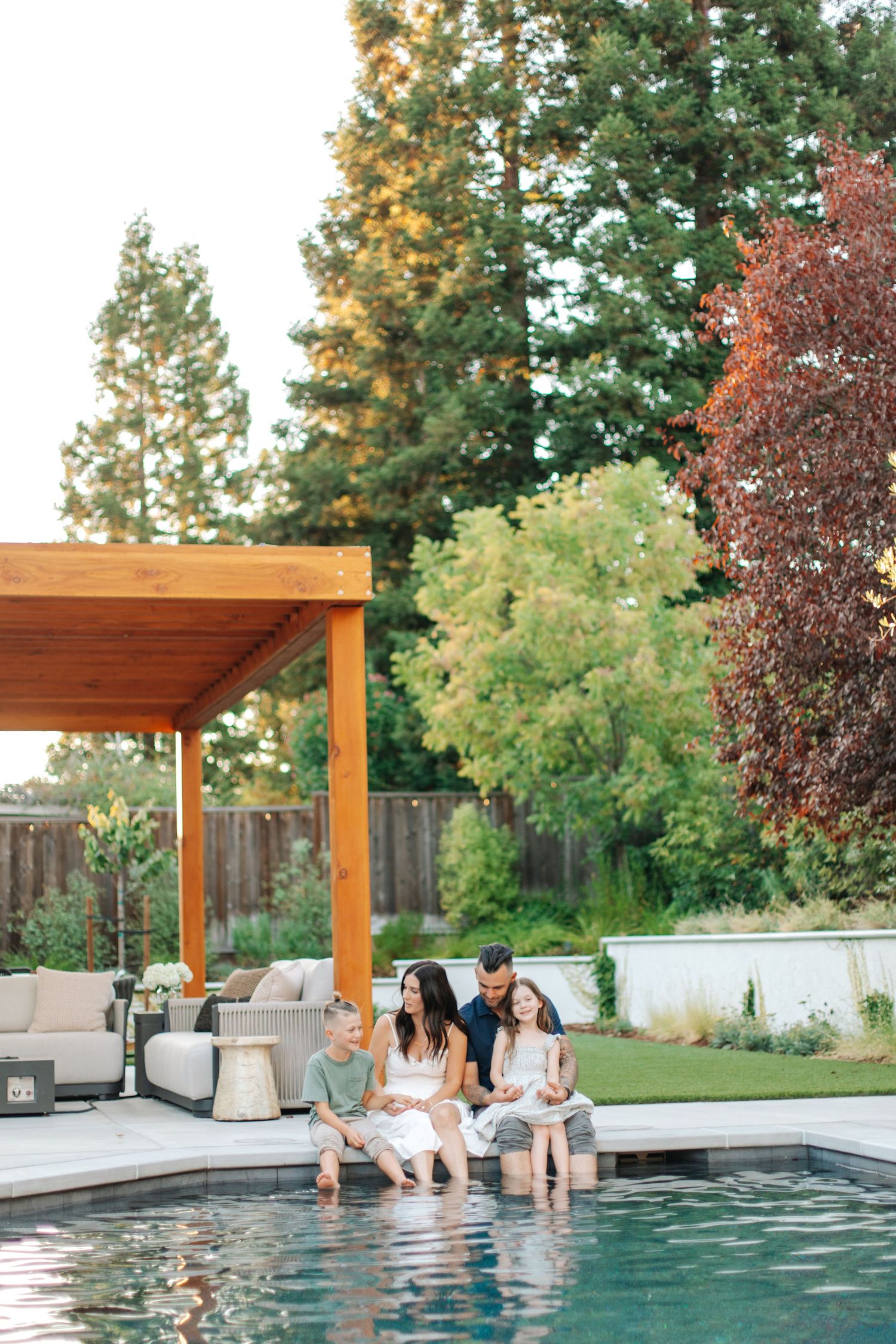 A family of four sits poolside at sunset, feet dangling in water beneath a wooden pergola.