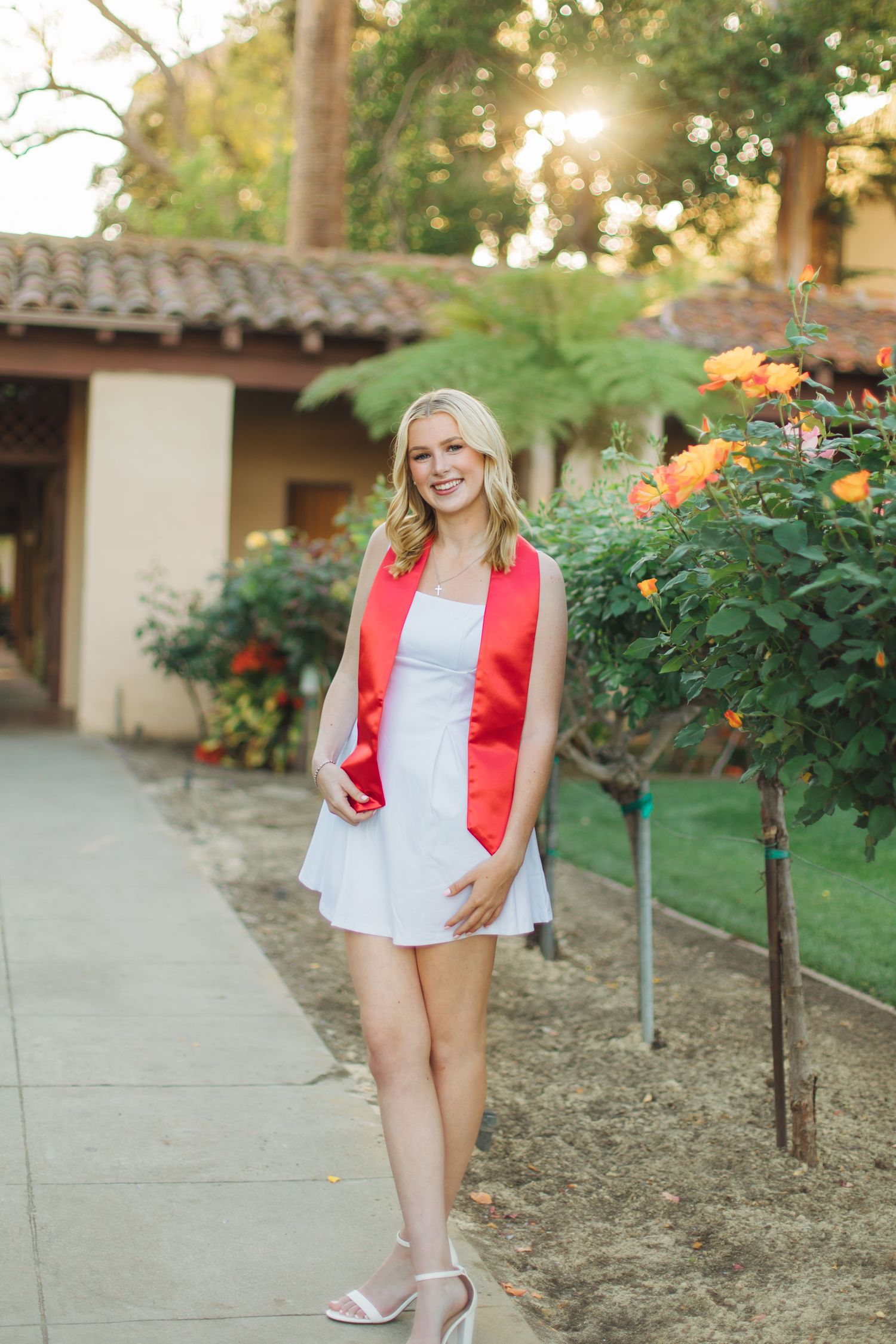 Blonde graduate in white dress and red stole poses on garden path with orange roses at sunset.