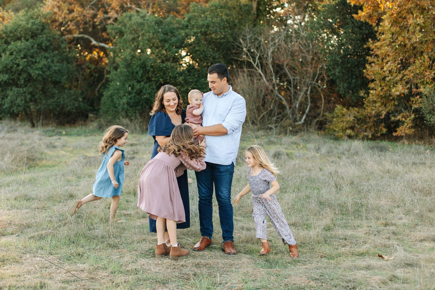 A cheerful family of five with three young daughters plays together in an open grassy field.