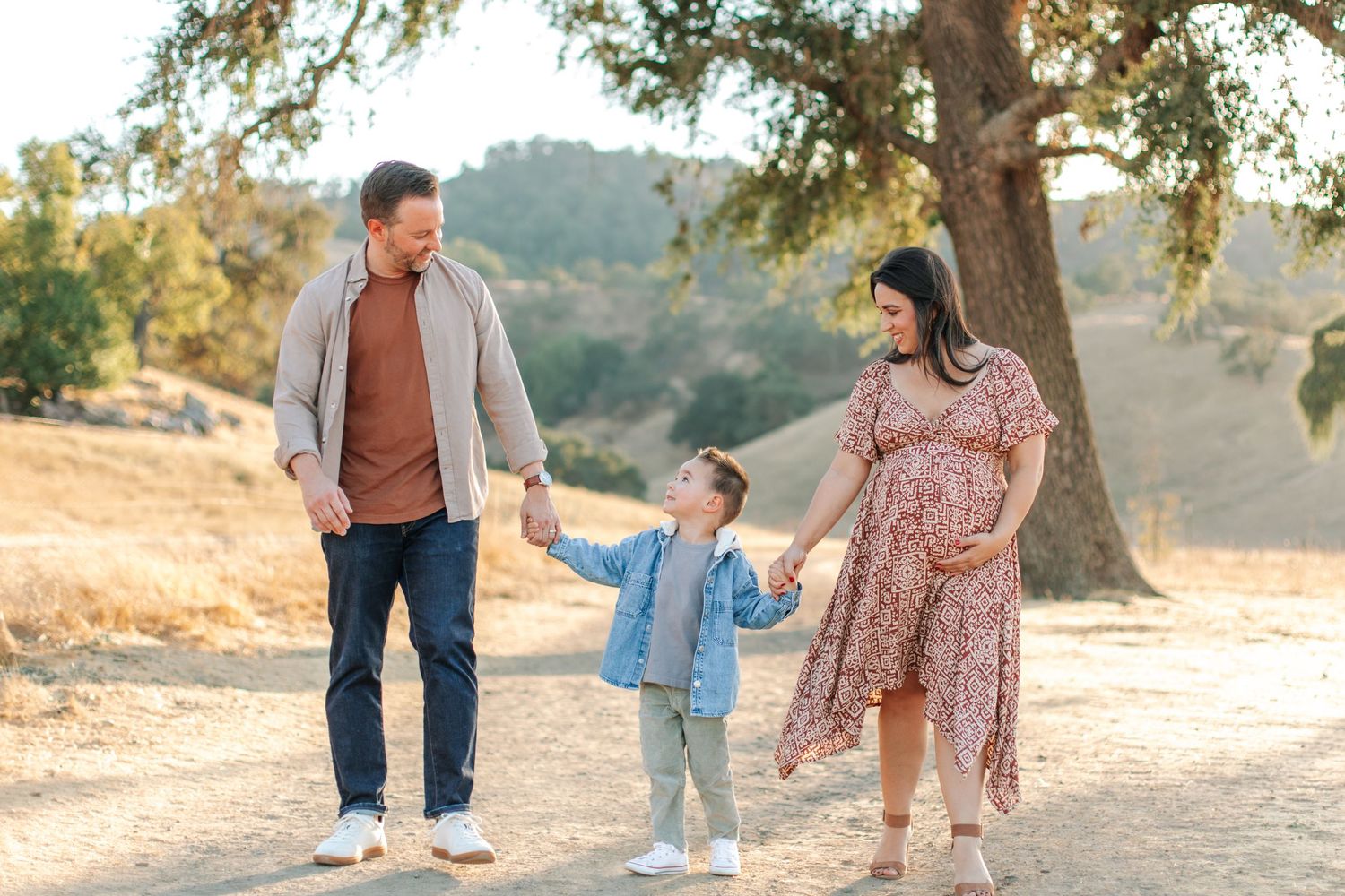 Pregnant woman in floral dress walks with husband and young son on a sunny trail.