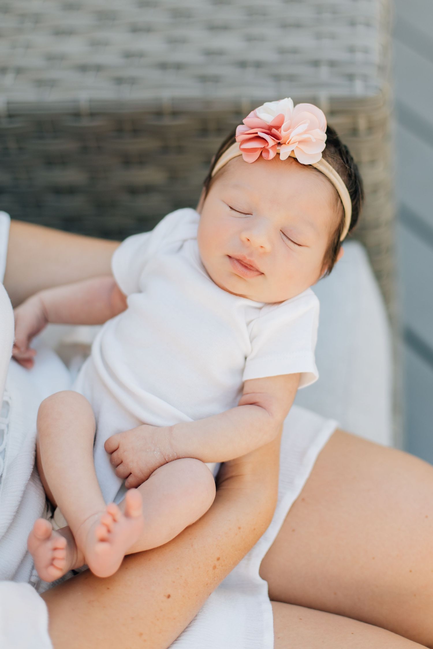 Peaceful sleeping newborn baby girl in white onesie with pink flower headband being held.