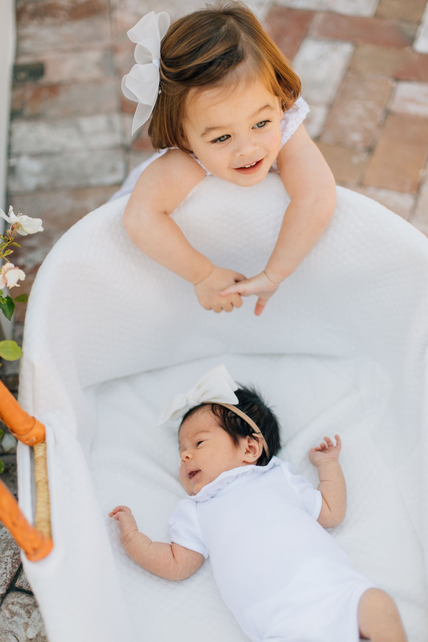 Toddler girl in white dress with bow smiles at newborn baby sibling lying in white bassinet.