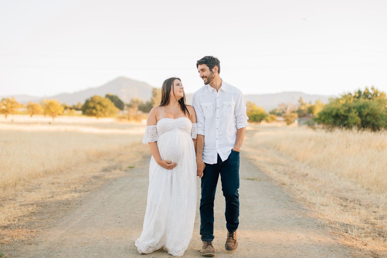 Expecting couple holding hands and smiling on rural dirt road with mountain backdrop at sunset.