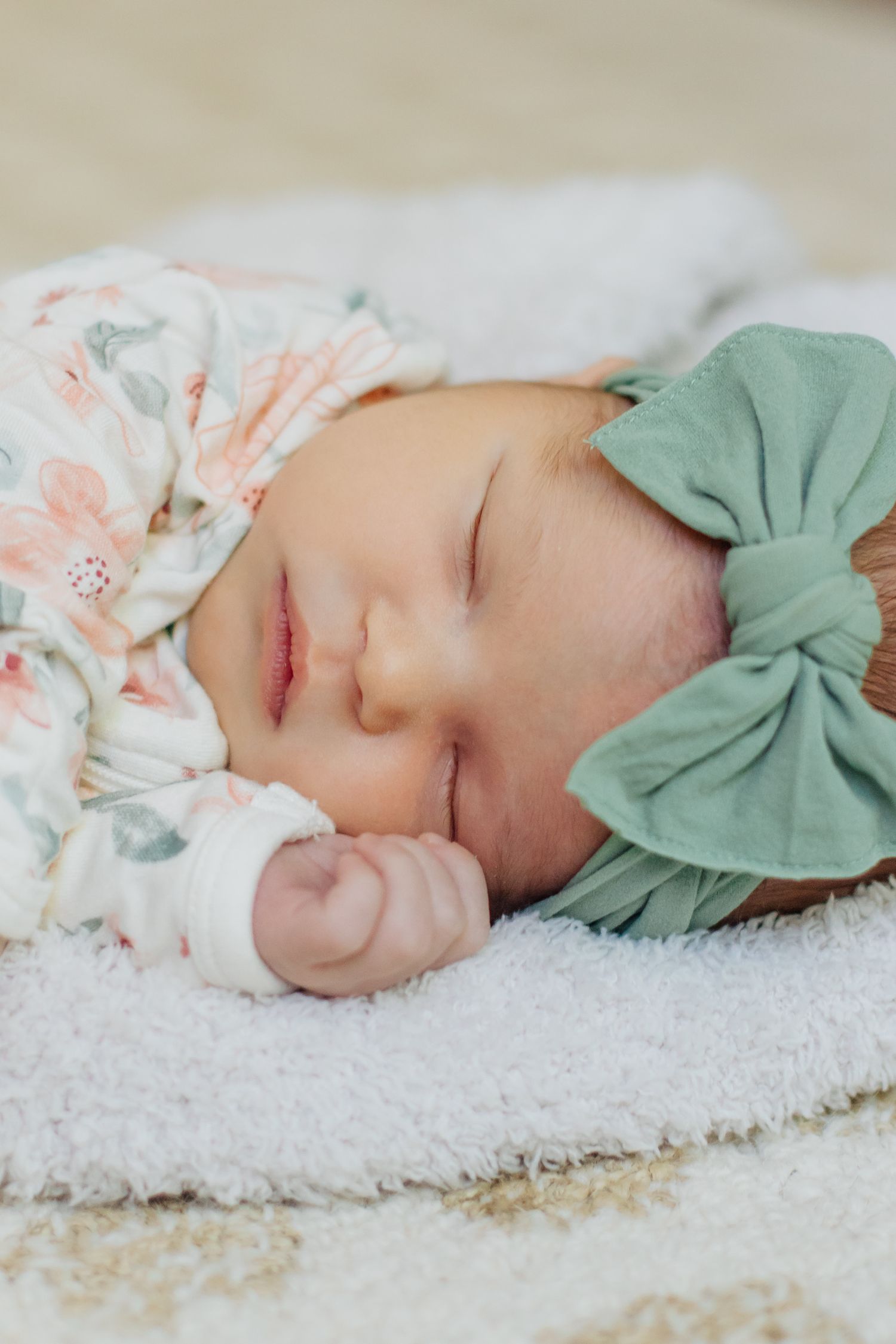Peaceful newborn baby girl sleeping on white blanket wearing green bow headband and floral outfit.