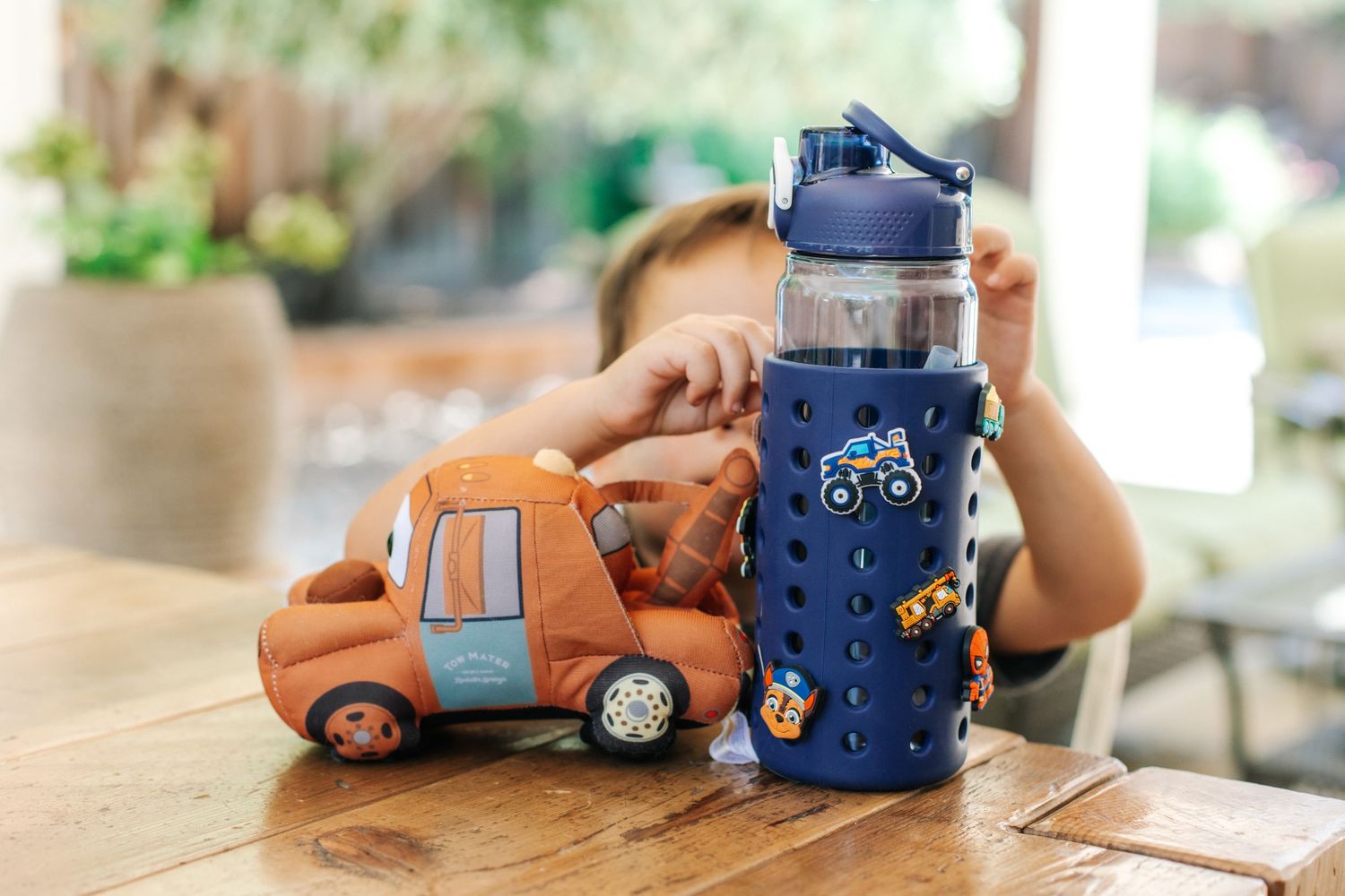Child playing with toy car next to navy blue character charm water bottle on wooden table.