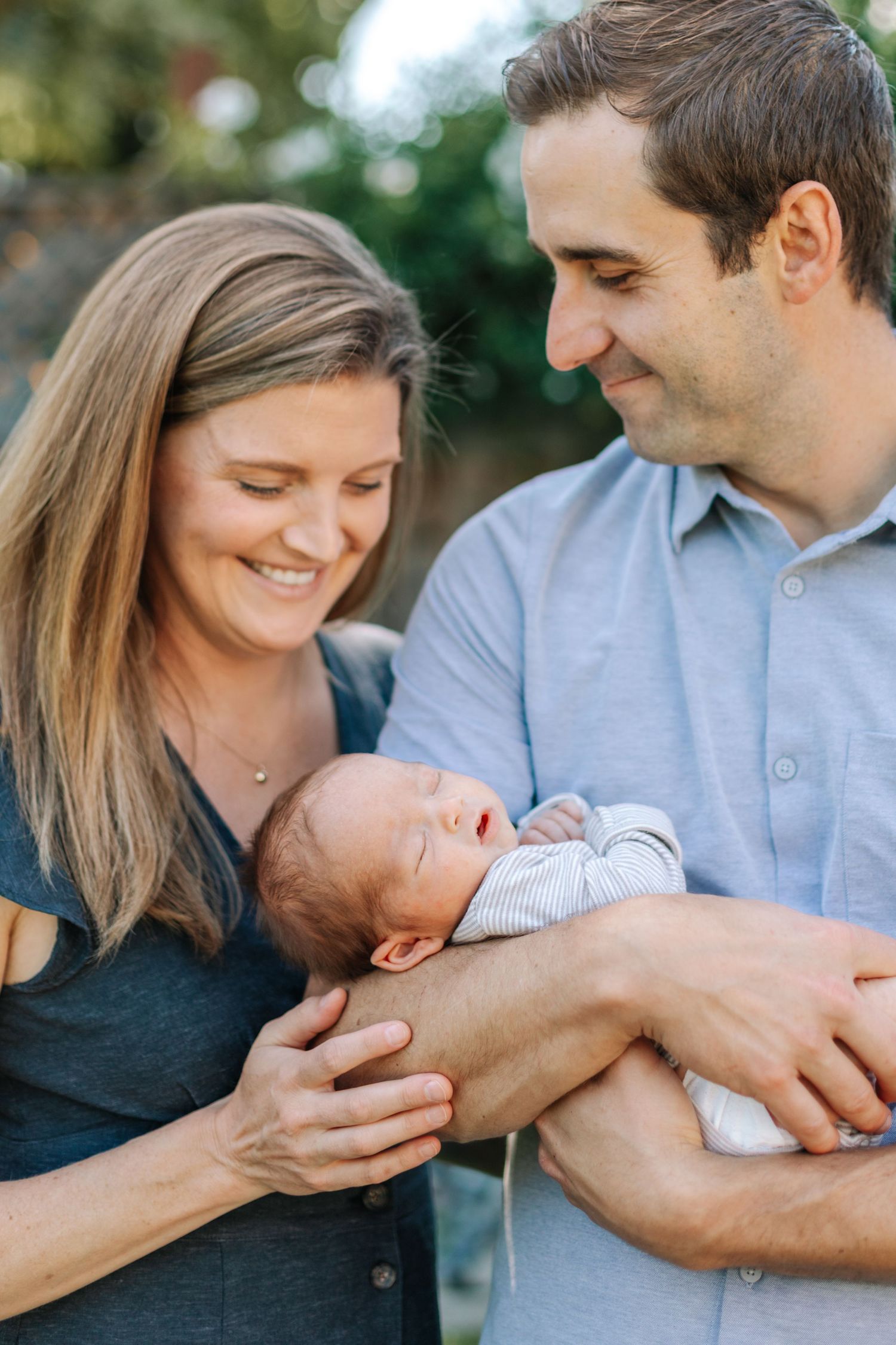 Smiling parents in outdoor garden setting gazing adoringly at their sleeping newborn baby together.