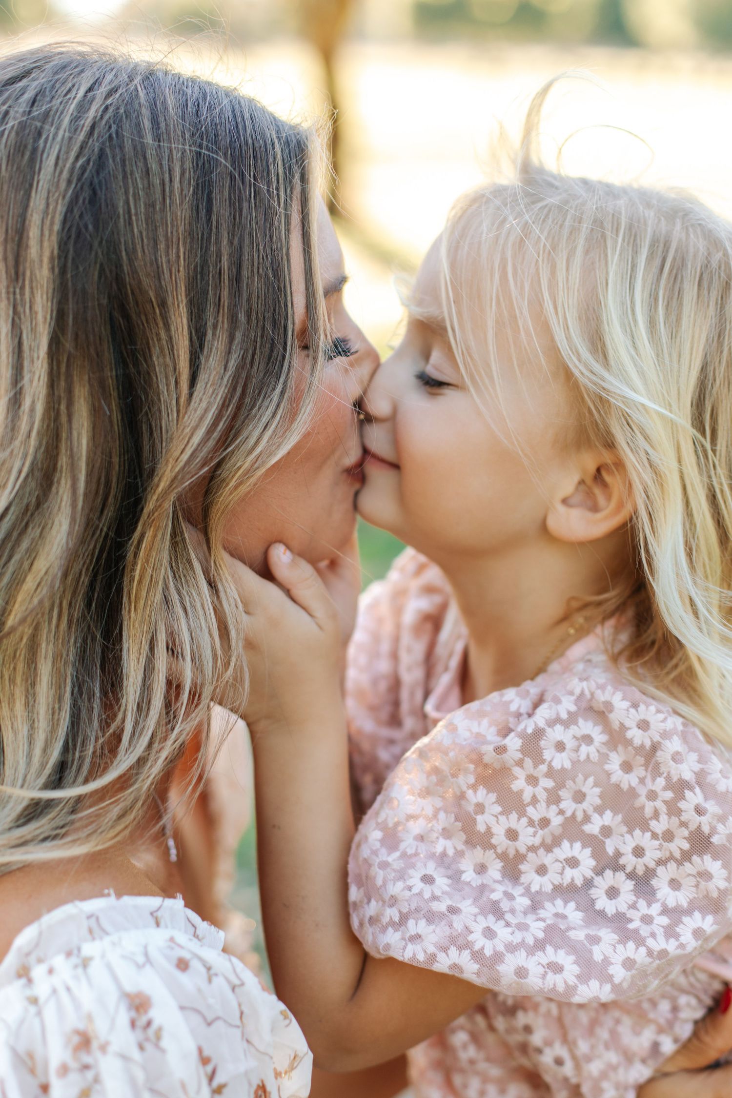 Sweet close-up of a mother and young blonde daughter sharing a tender kiss outdoors in warm sunlight.