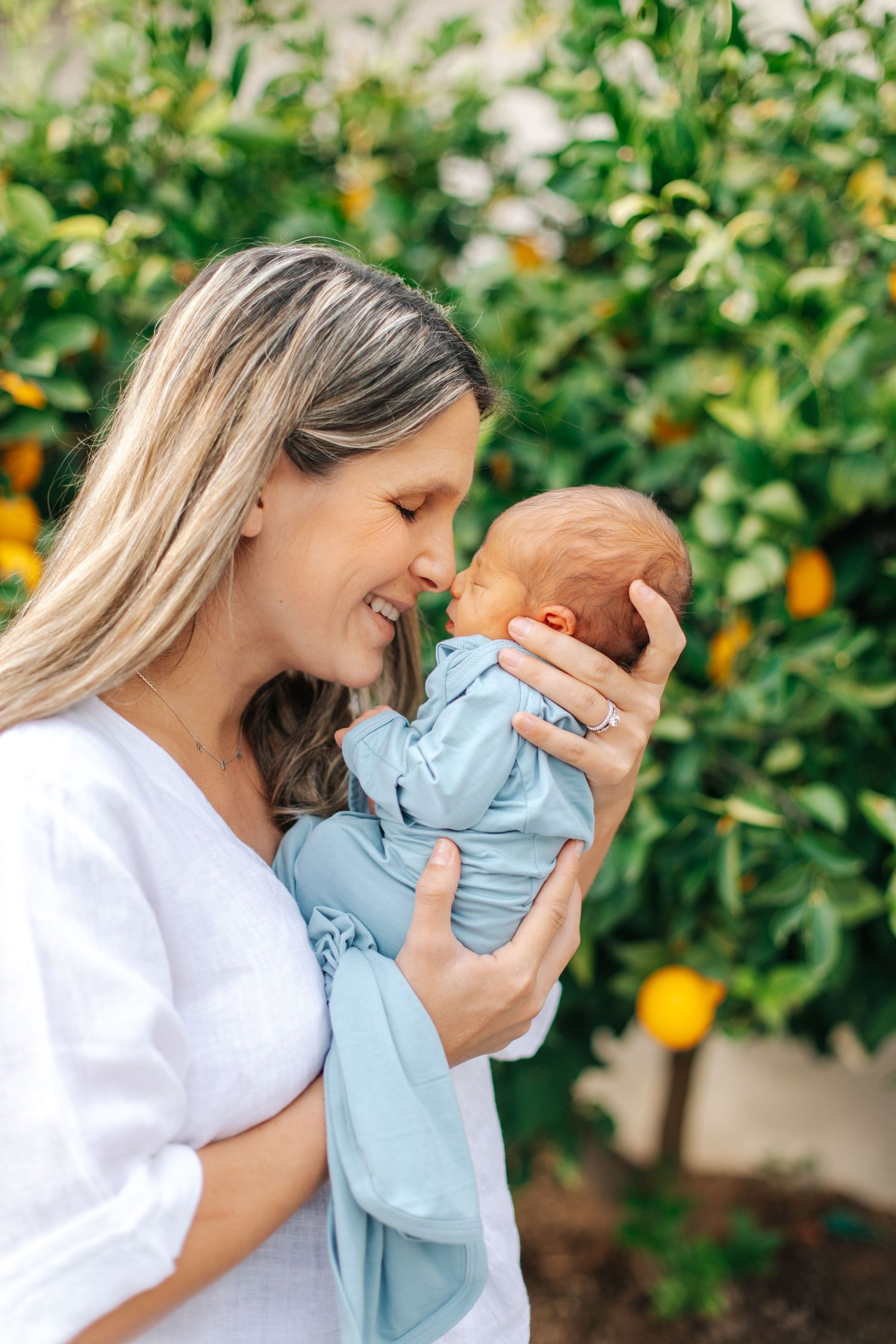 Blonde mother smiling and nuzzling her newborn baby wrapped in blue swaddle outdoors.