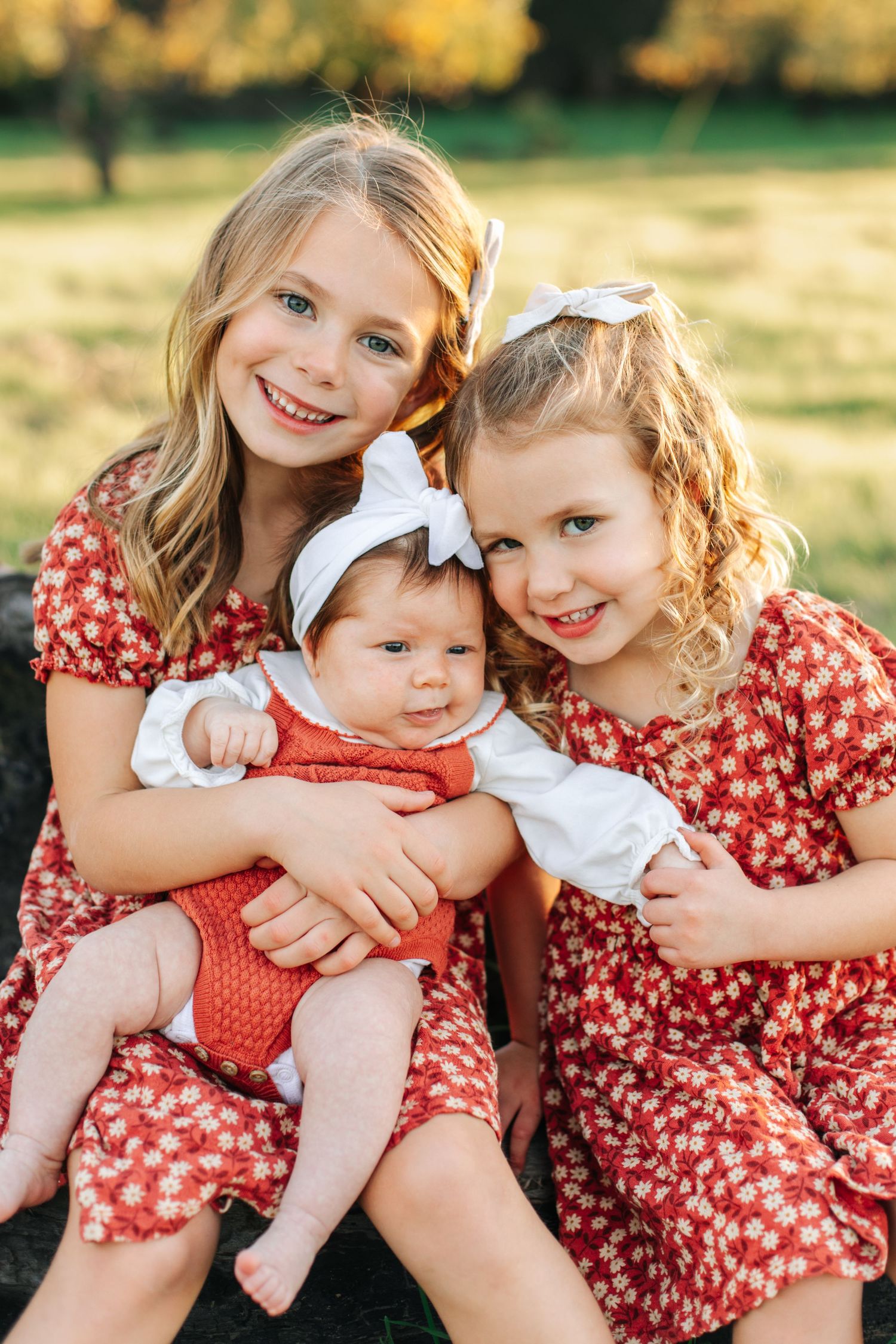 Two young sisters in red floral dresses lovingly hold their newborn baby sibling outdoors.