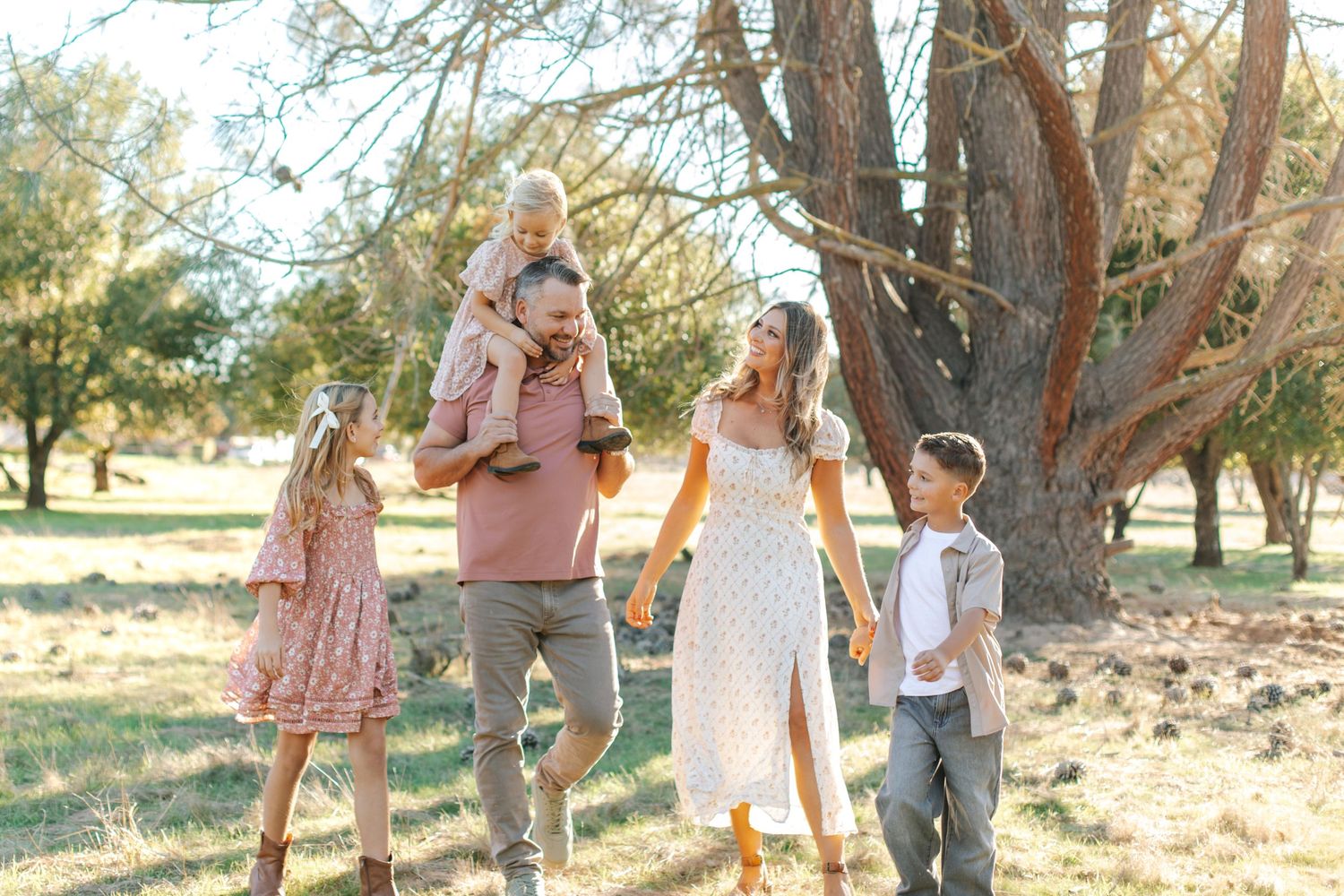 Active family of four walking and laughing together in a sunny park with large trees.