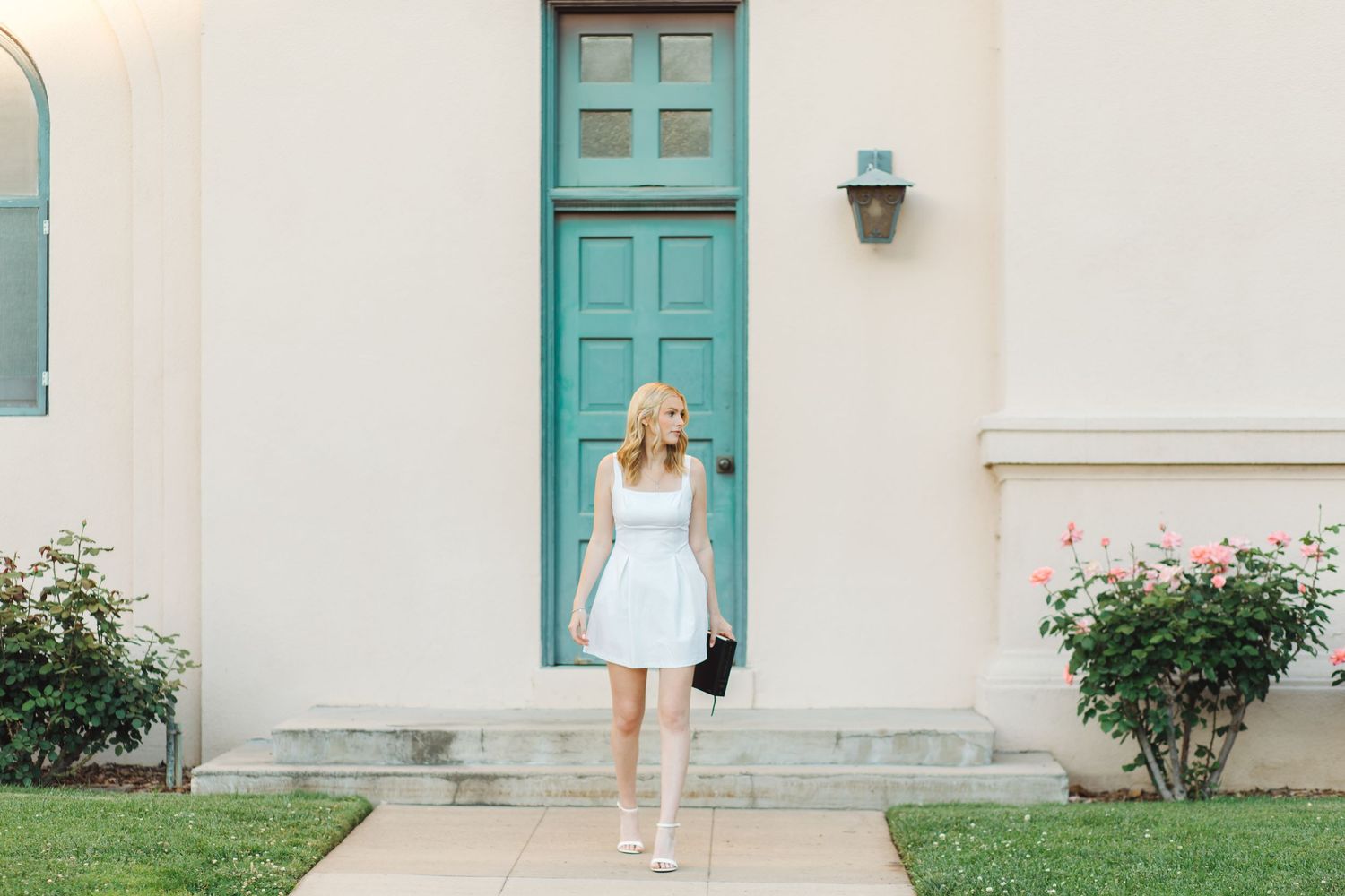 Blonde woman in white dress walking toward teal door of a light-colored building with roses.