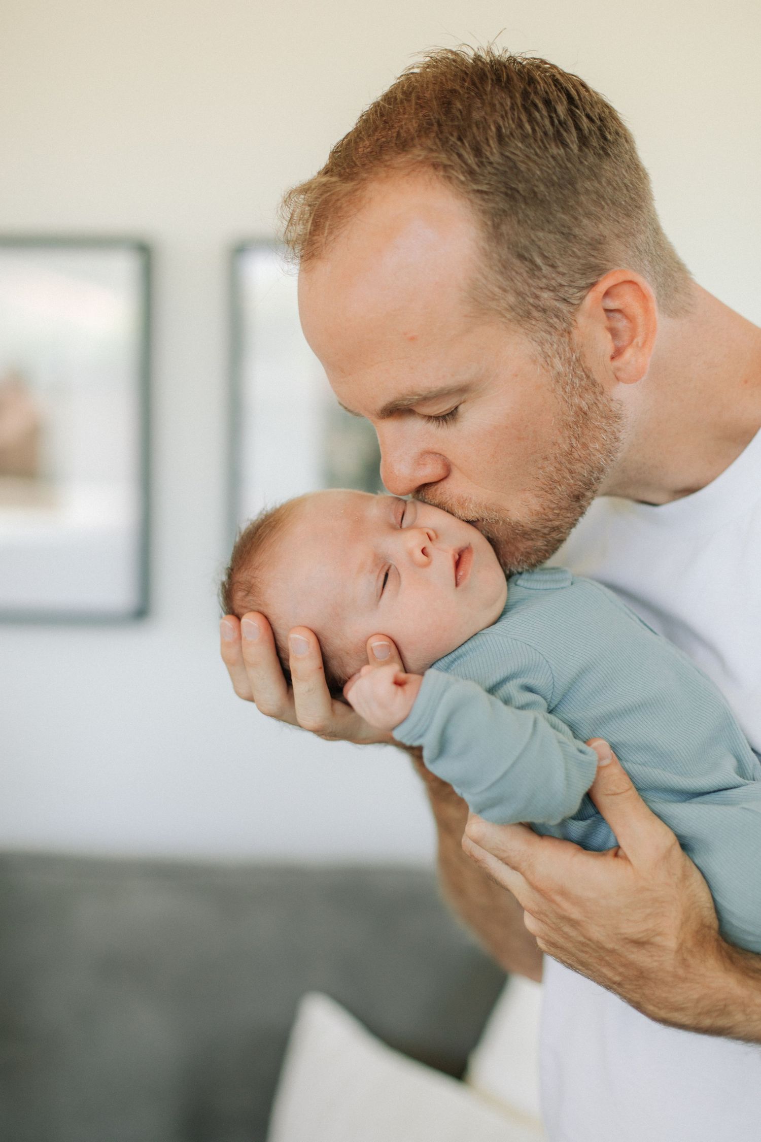 A father gently kissing his sleeping newborn baby dressed in a light blue onesie at home.