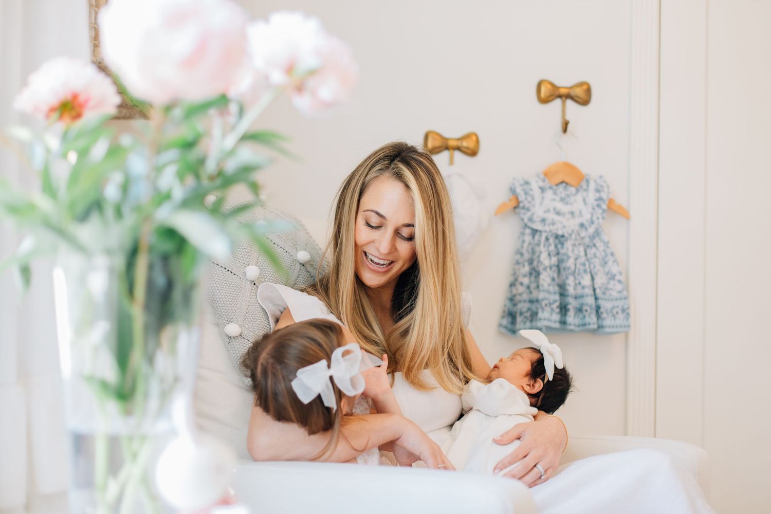 Joyful mother smiles at toddler daughter and newborn baby on a white bed in bright nursery.