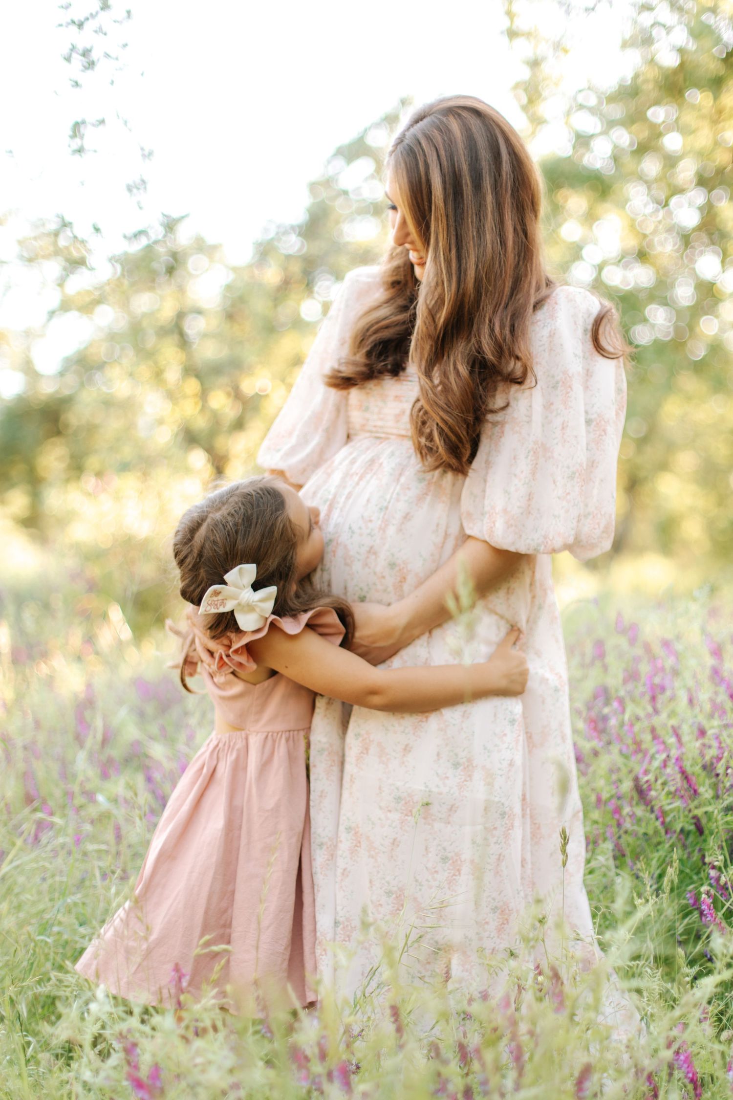 Young girl in pink dress hugging her pregnant mothers belly in a beautiful wildflower meadow.