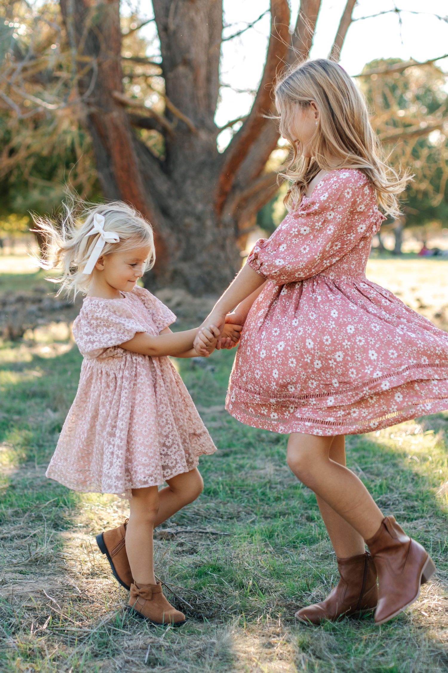 Two young girls in pink dresses hold hands and spin together near a large tree outdoors.