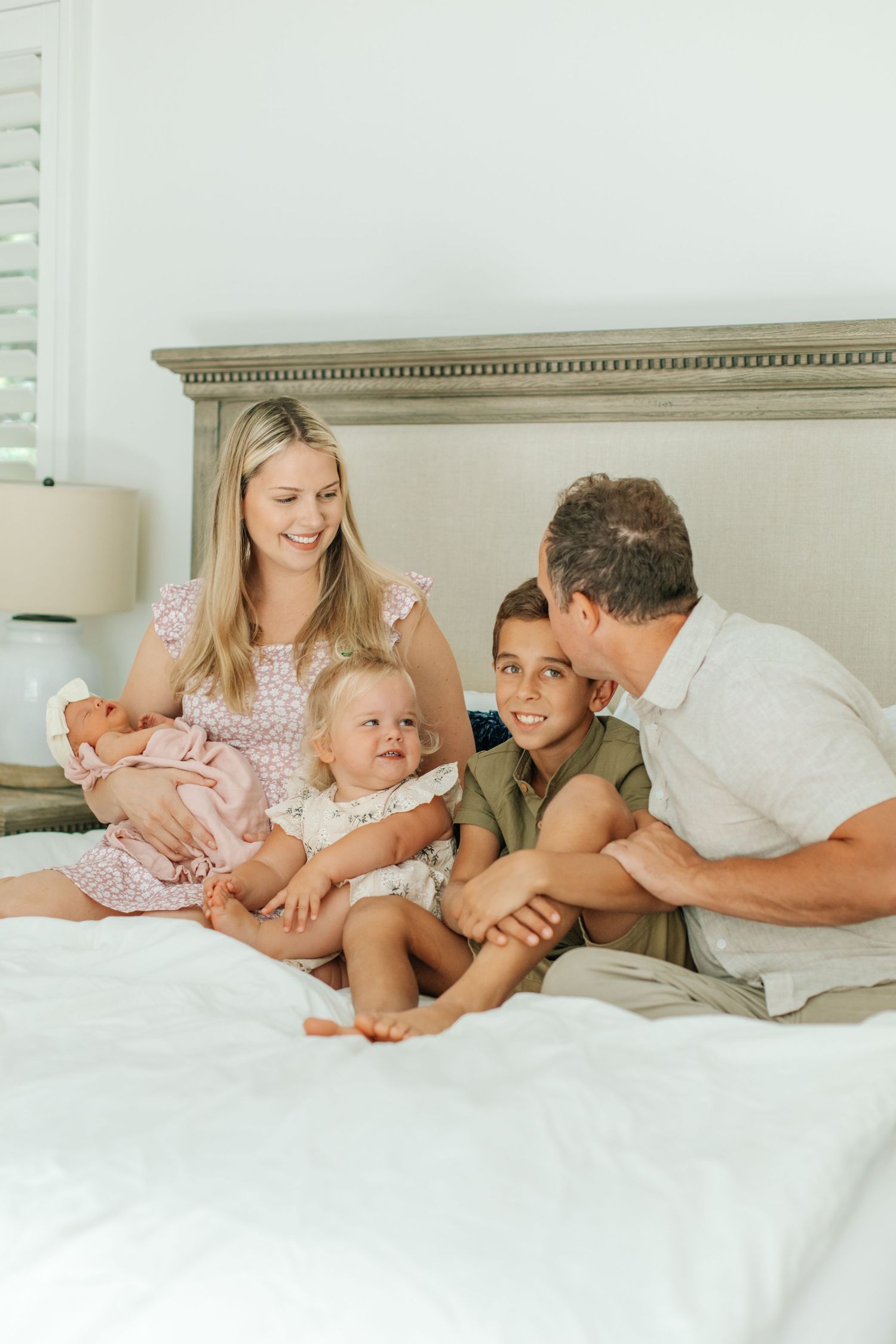 Happy family sits on a bed smiling, mother holding newborn baby girl surrounded by two older children.
