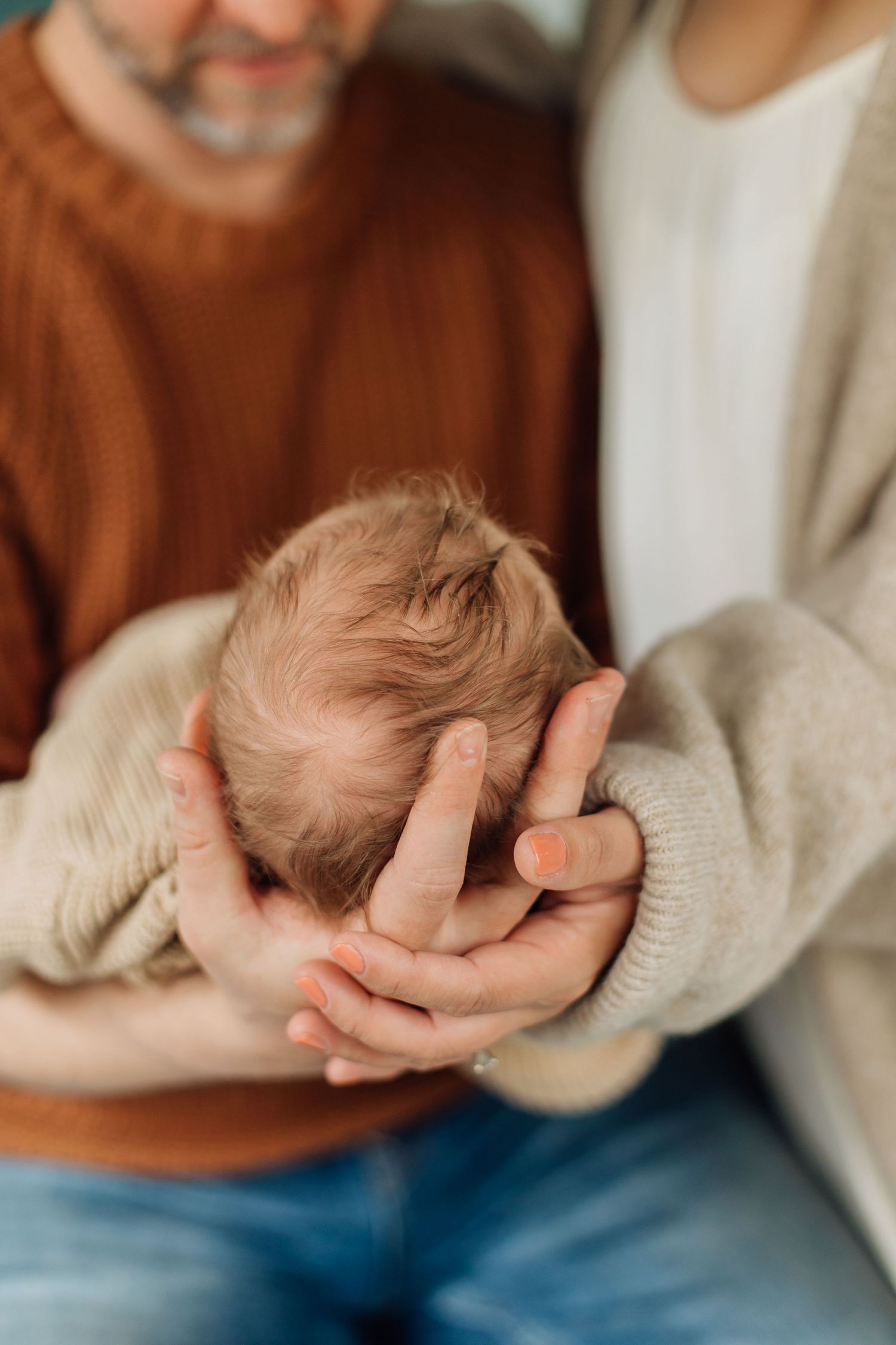 Parents cradling newborn baby's head in their hands, wearing brown and cream sweaters.