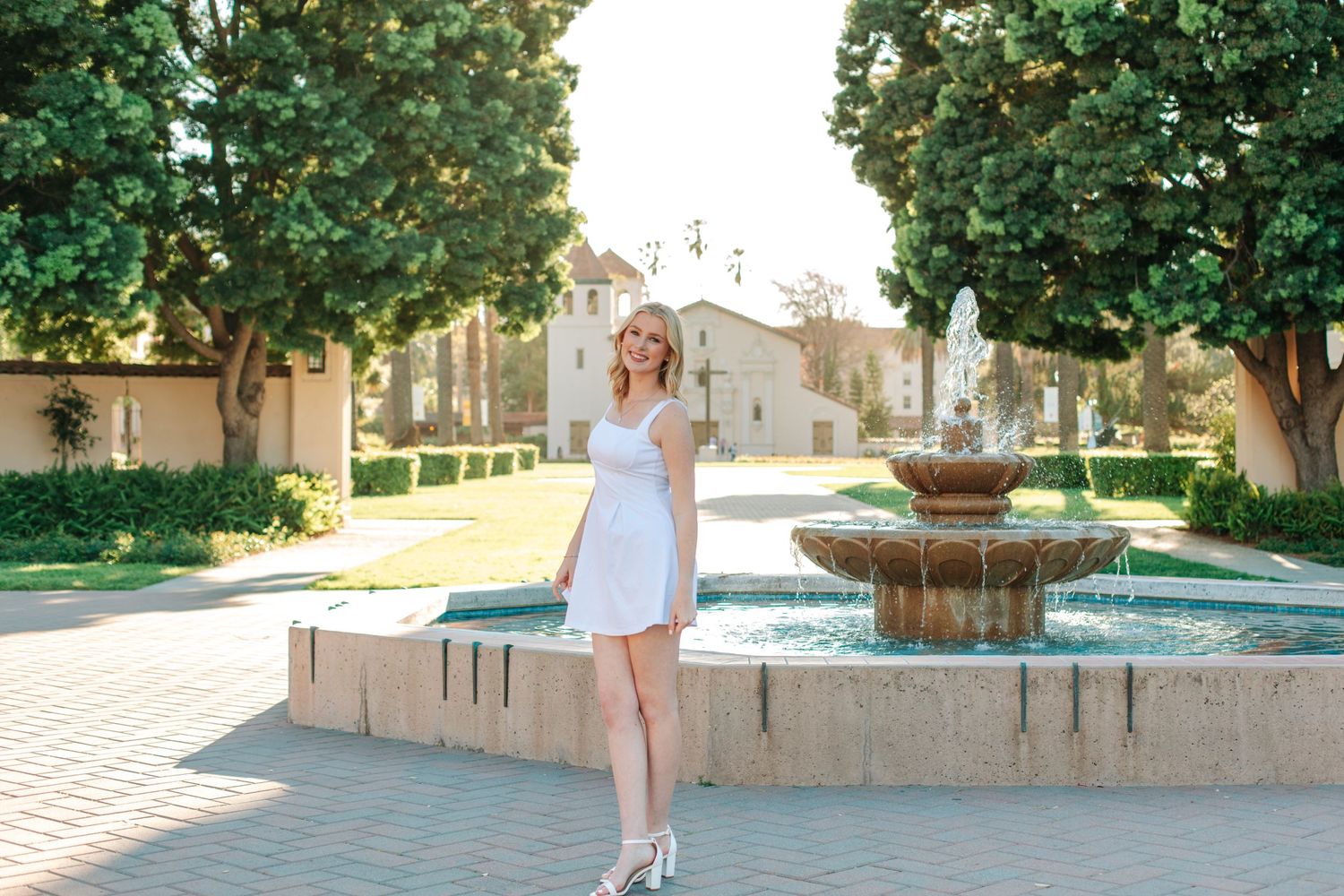 A blonde young woman in a white dress poses near a decorative fountain on a sunny day.
