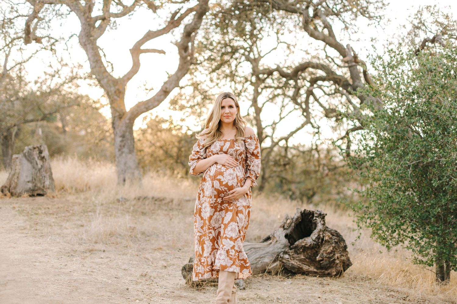 Pregnant woman in rust floral maxi dress standing among oak trees in a golden outdoor setting.