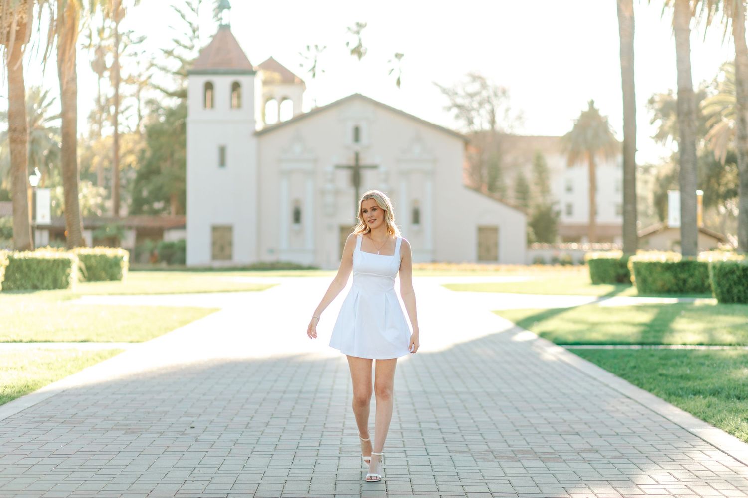 Blonde woman in white dress walking toward camera on brick path with white mission-style church backdrop.