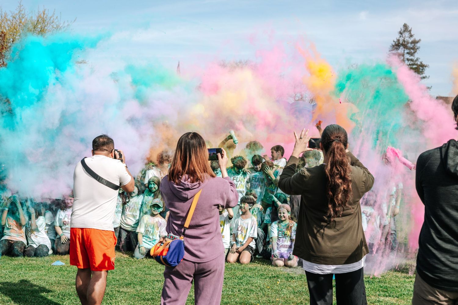 Crowd throwing vibrant multicolored powder at outdoor fun run celebration event on sunny day.