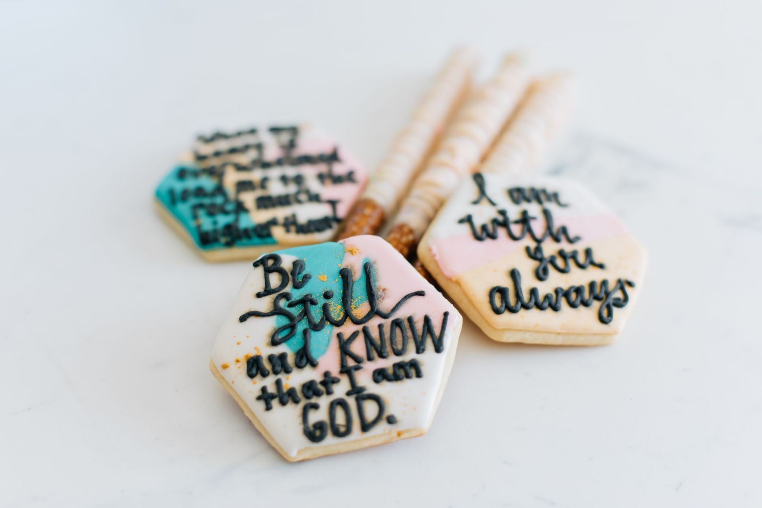 Hexagonal scripture-quote sugar cookies with pastel watercolor frosting and decorative striped sticks.