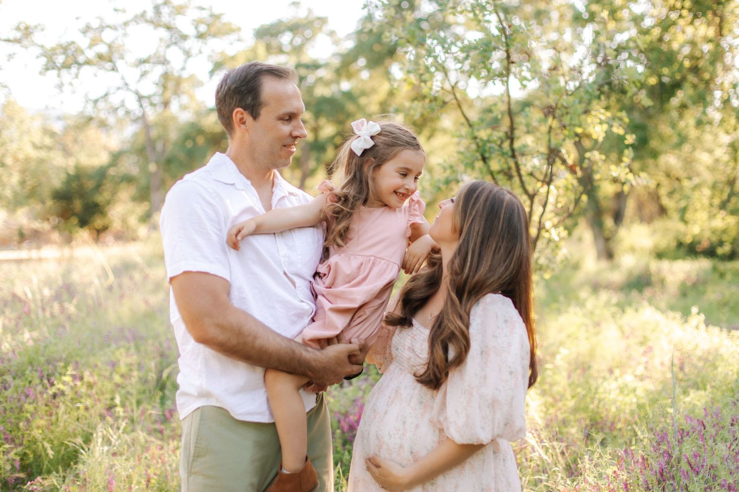 Happy family with pregnant mother, father, and young daughter in a sunny wildflower meadow.