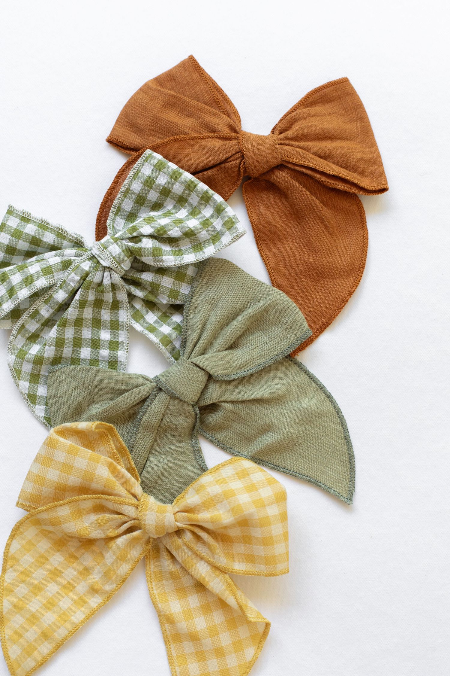 Four fabric hair bows in brown, green gingham, sage, and yellow gingham patterns on white background.