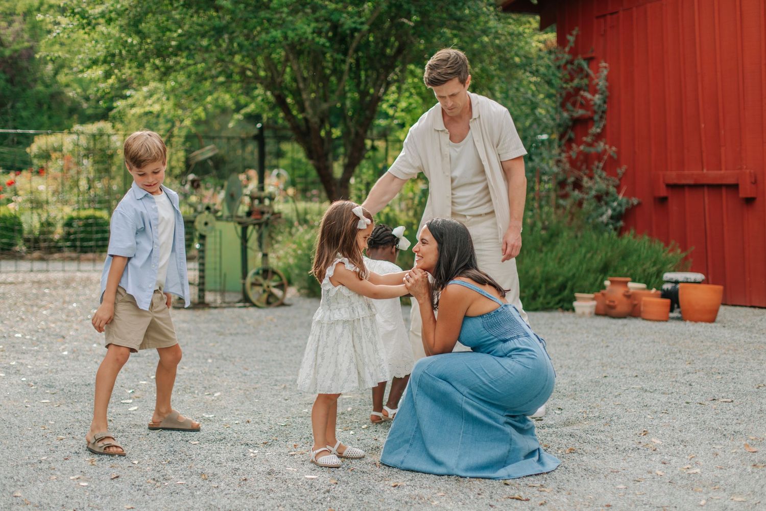 Family of four enjoying a sunny outdoor moment near a red barn, with parents interacting warmly with young children.