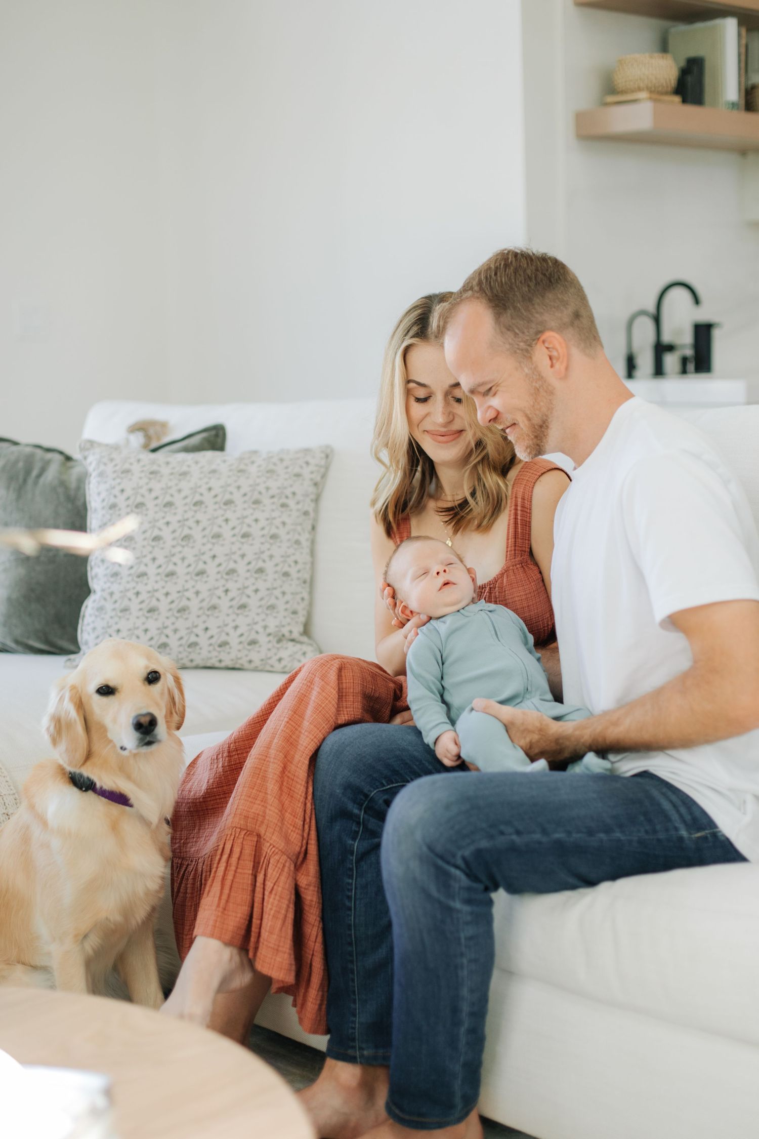 Loving parents admiring their sleeping newborn baby on a white couch with a golden retriever nearby.