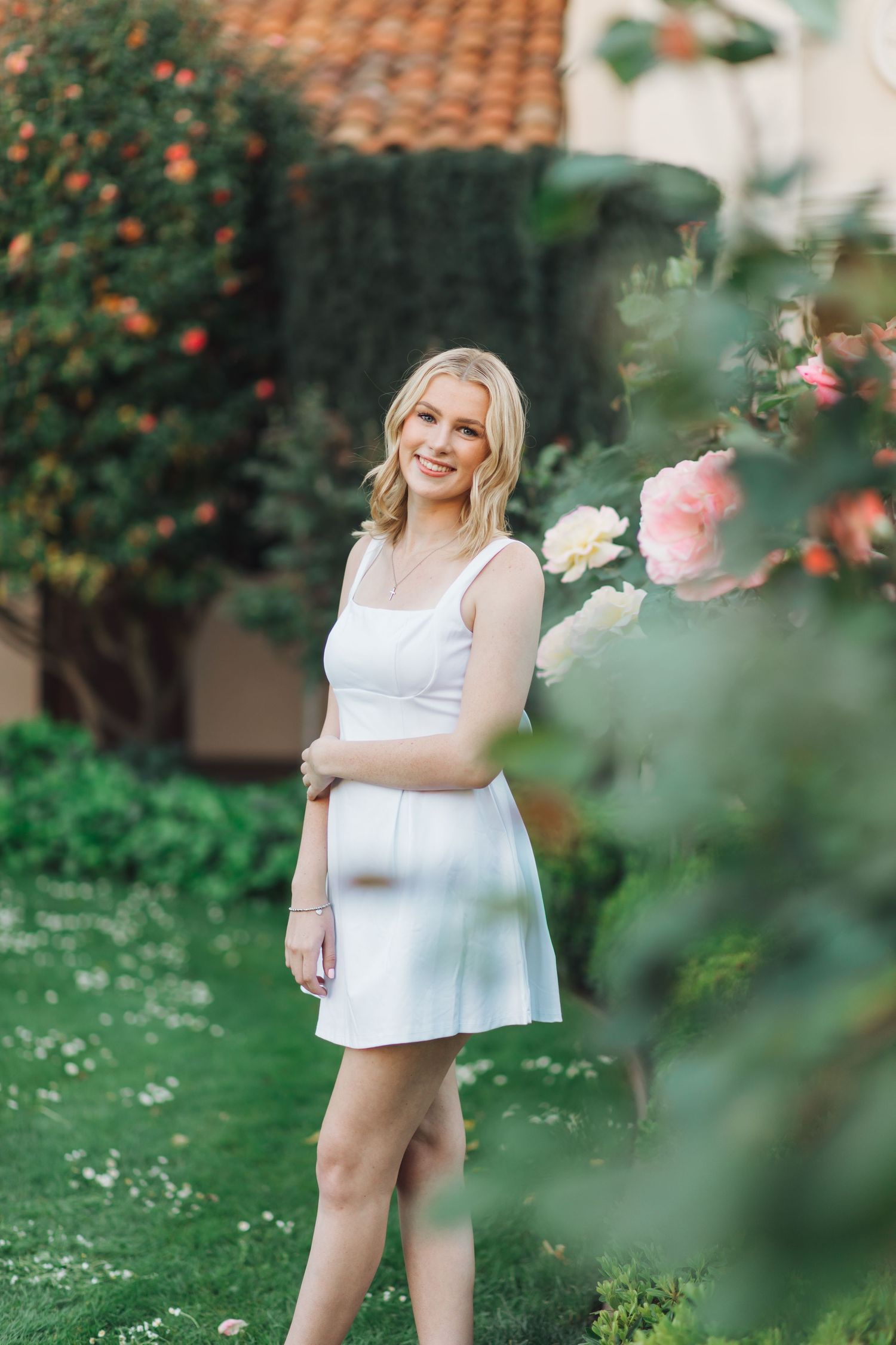 Smiling blonde woman in white sundress posing among pink roses and greenery in a garden.