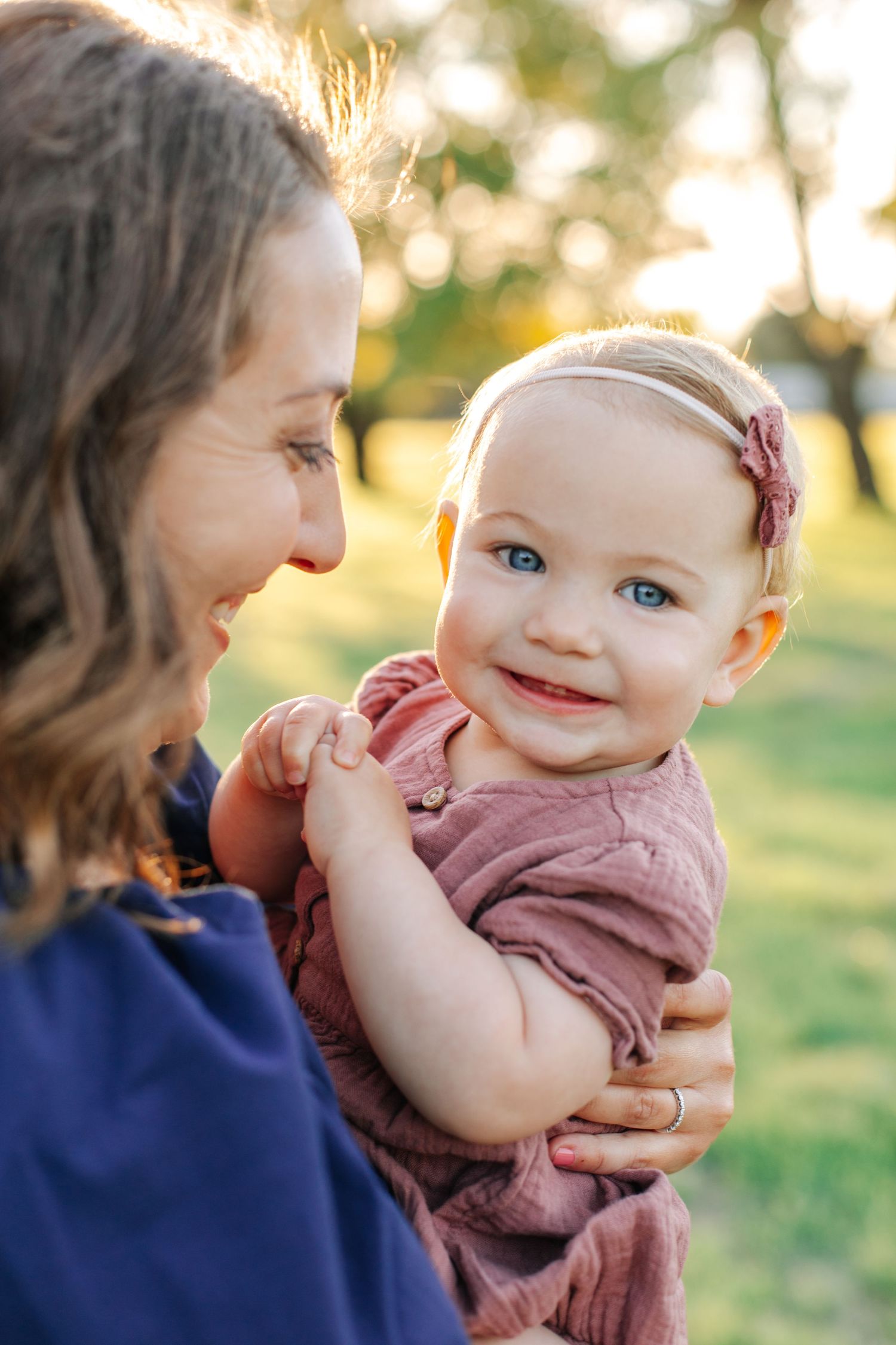 A smiling blue-eyed baby girl in a pink outfit is held lovingly by her mother outdoors.
