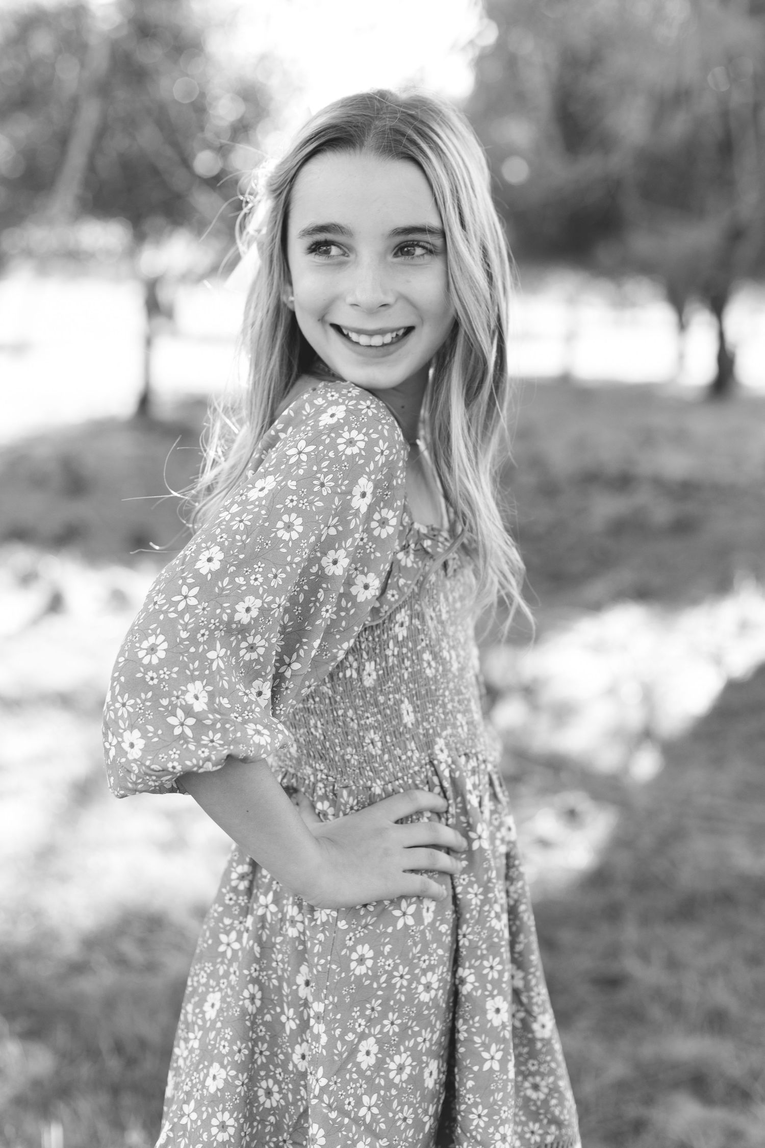 A smiling blonde girl in a floral dress poses confidently in a black and white outdoor portrait.
