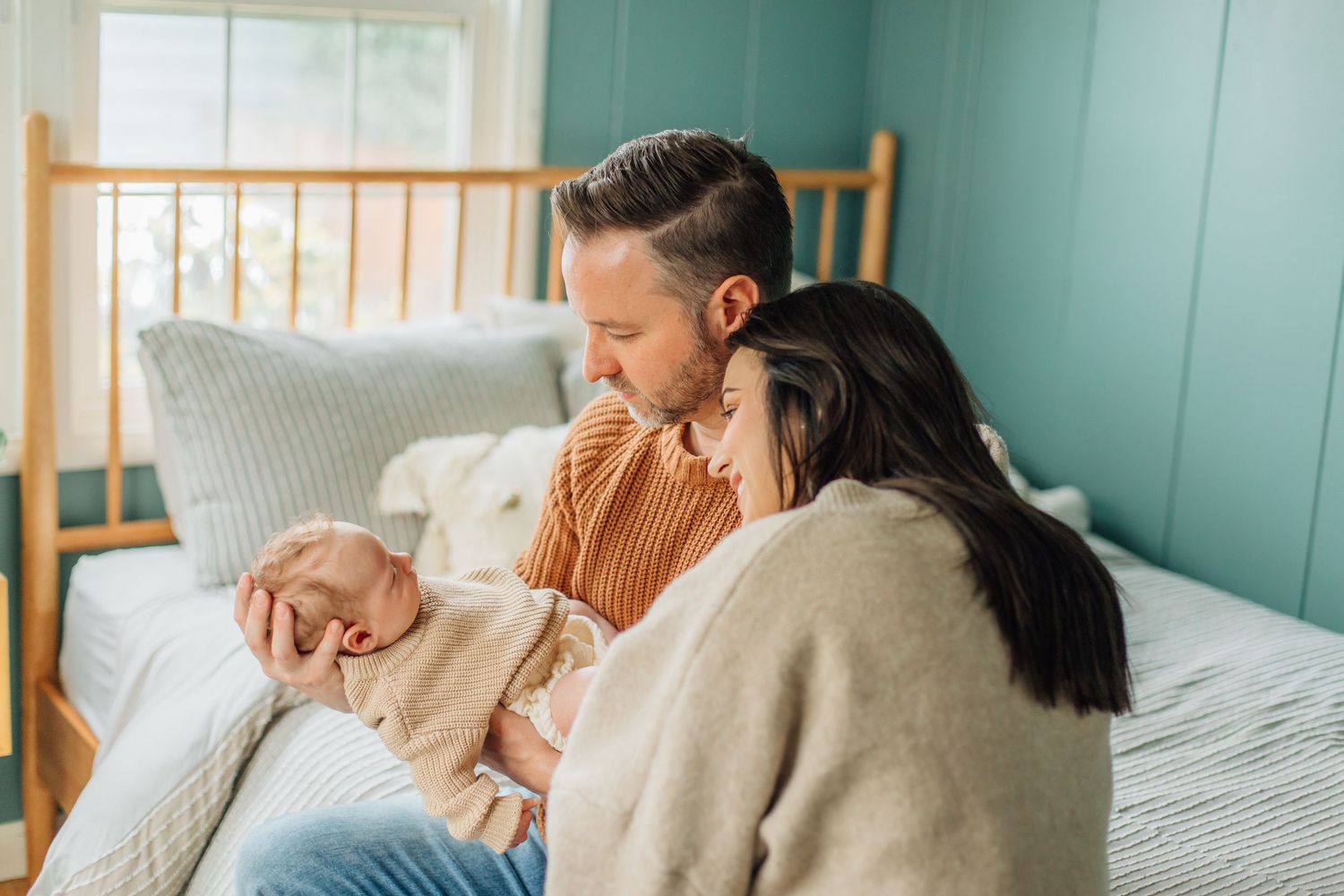 Happy couple sitting by bed admiring their newborn baby in warm natural light.
