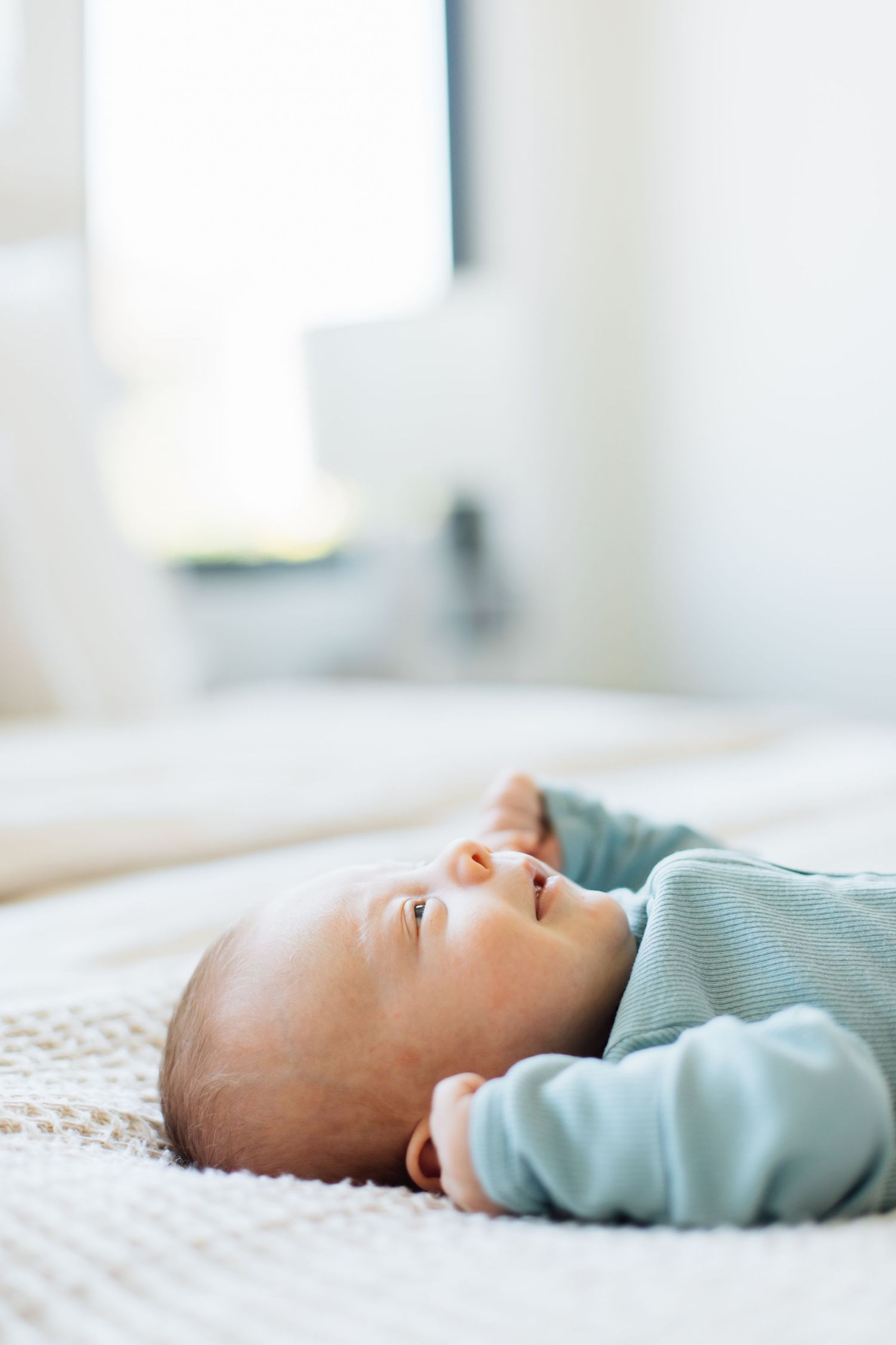 Peaceful newborn baby in blue striped onesie lying on white bed gazing upward in bright airy room.