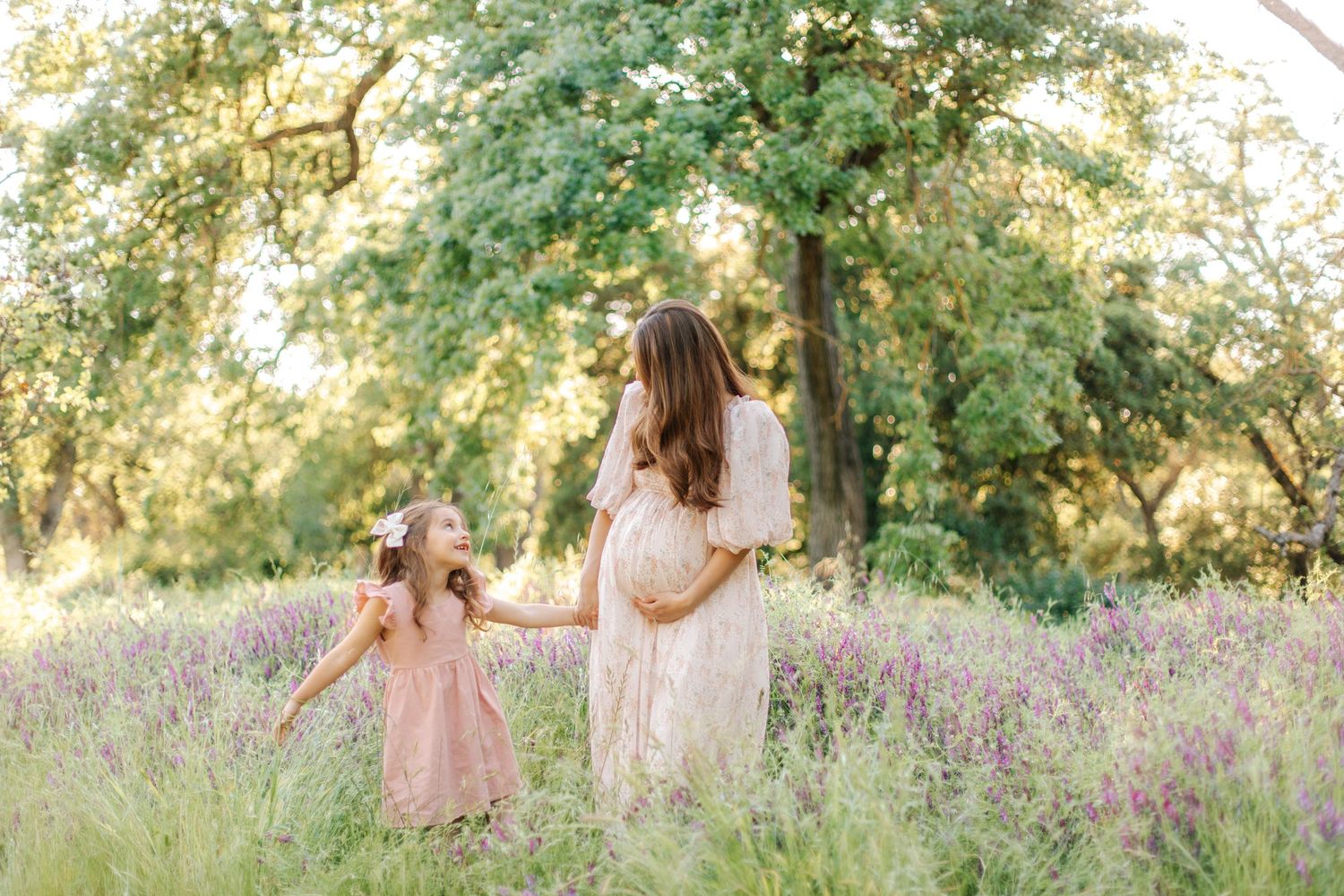 Pregnant mother and young daughter holding hands walking through a sunny lavender wildflower field.