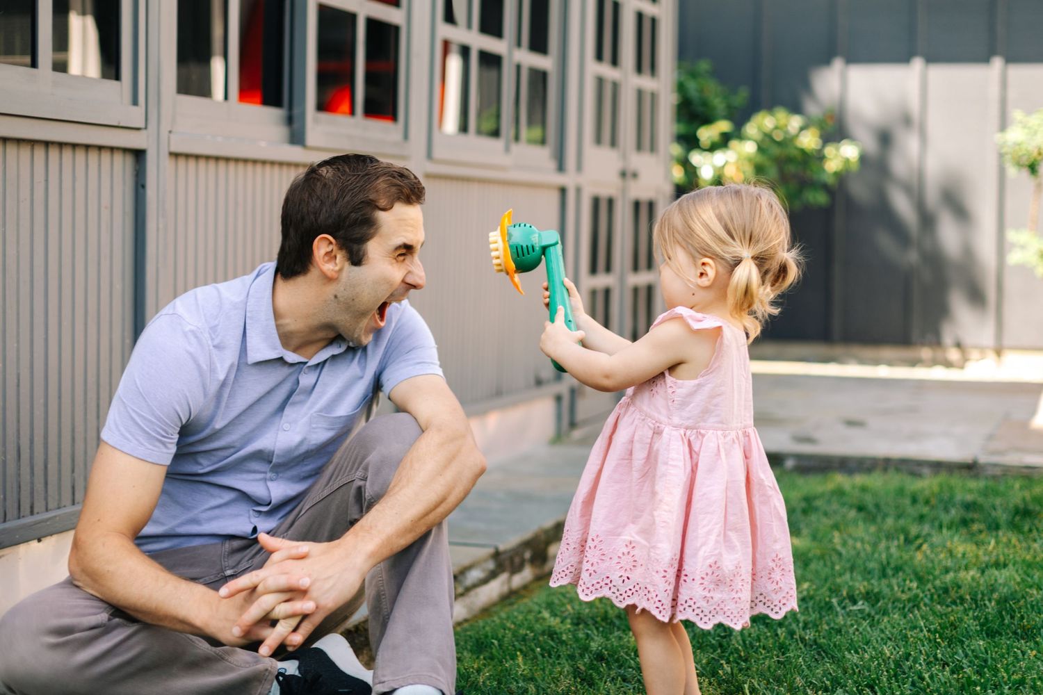 A toddler girl points a bubble toy at her laughing father in a backyard.