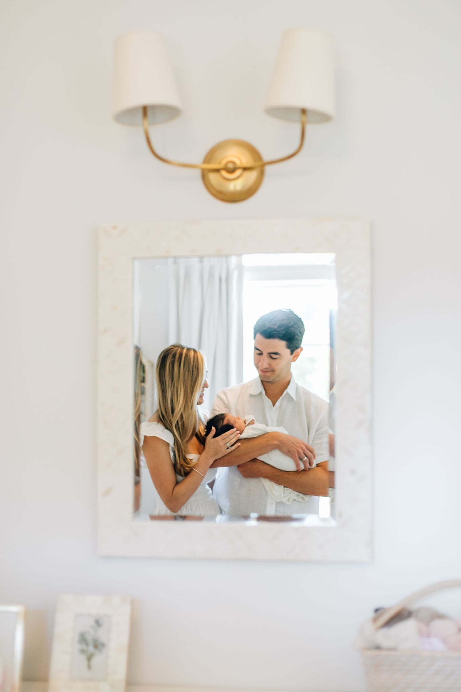 Couple with newborn baby reflected in an ornate white mirror with gold wall sconce above.