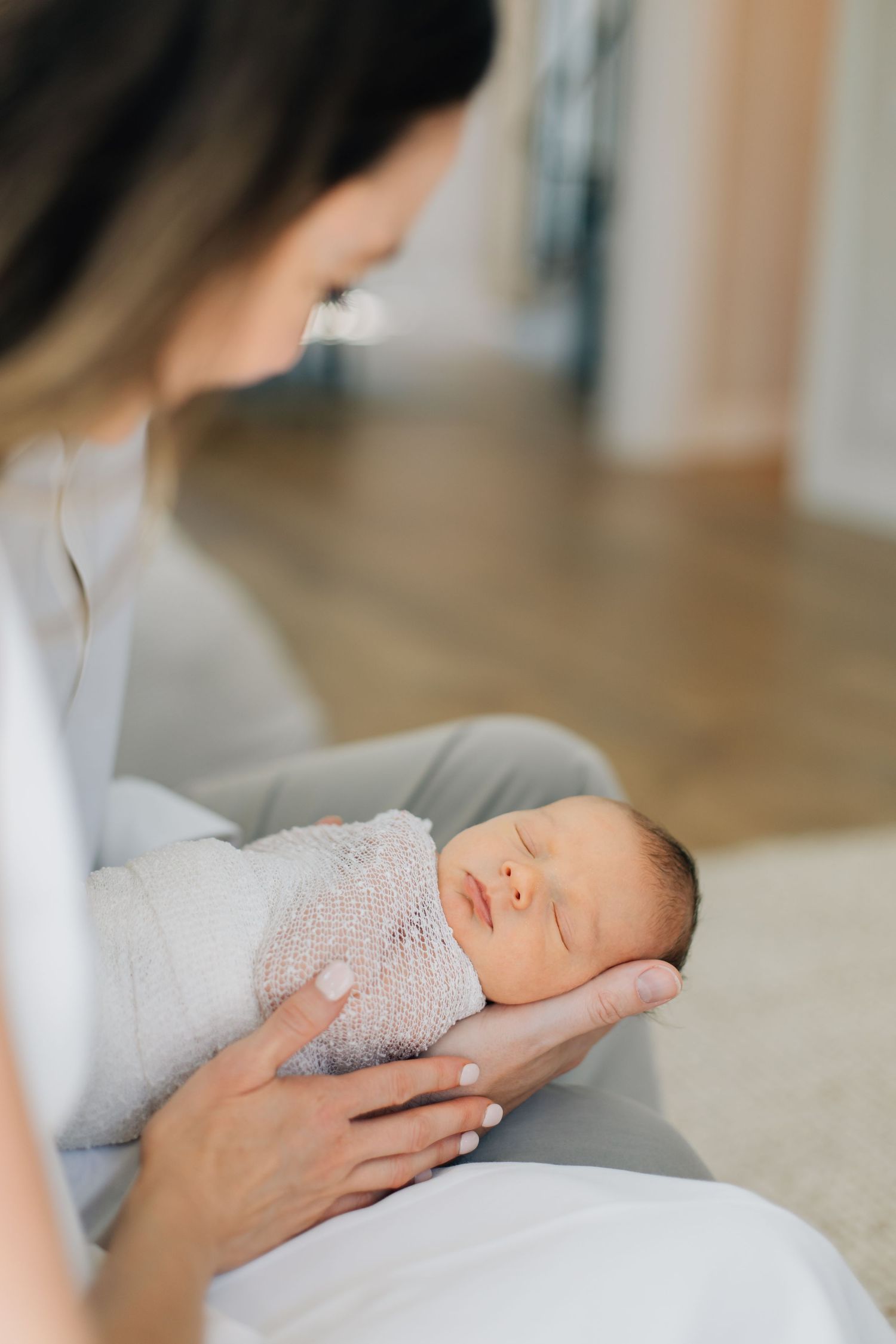 Mother tenderly holds her swaddled sleeping newborn baby in her arms indoors.