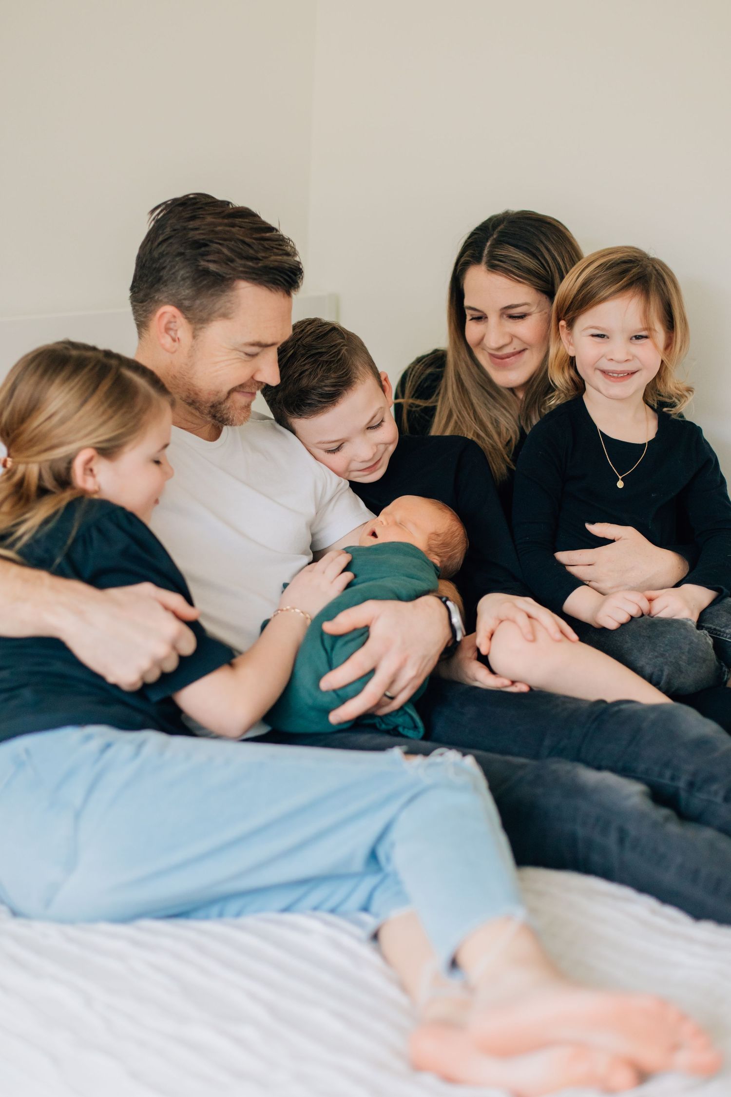 Happy family of six cuddles together on a bed admiring their newborn baby wrapped in teal.