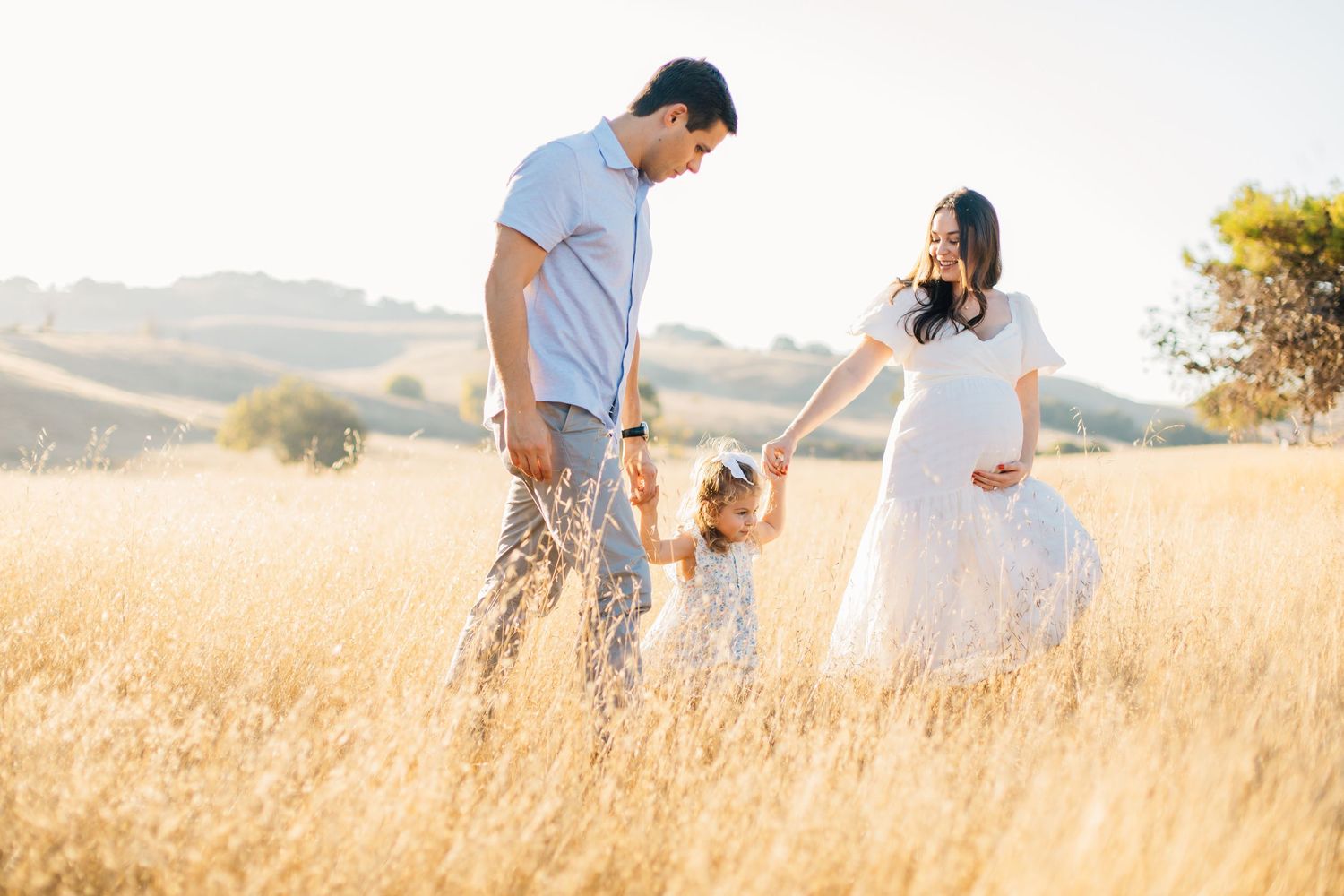 Family of three with pregnant mother holding hands with toddler and partner in golden grass field.