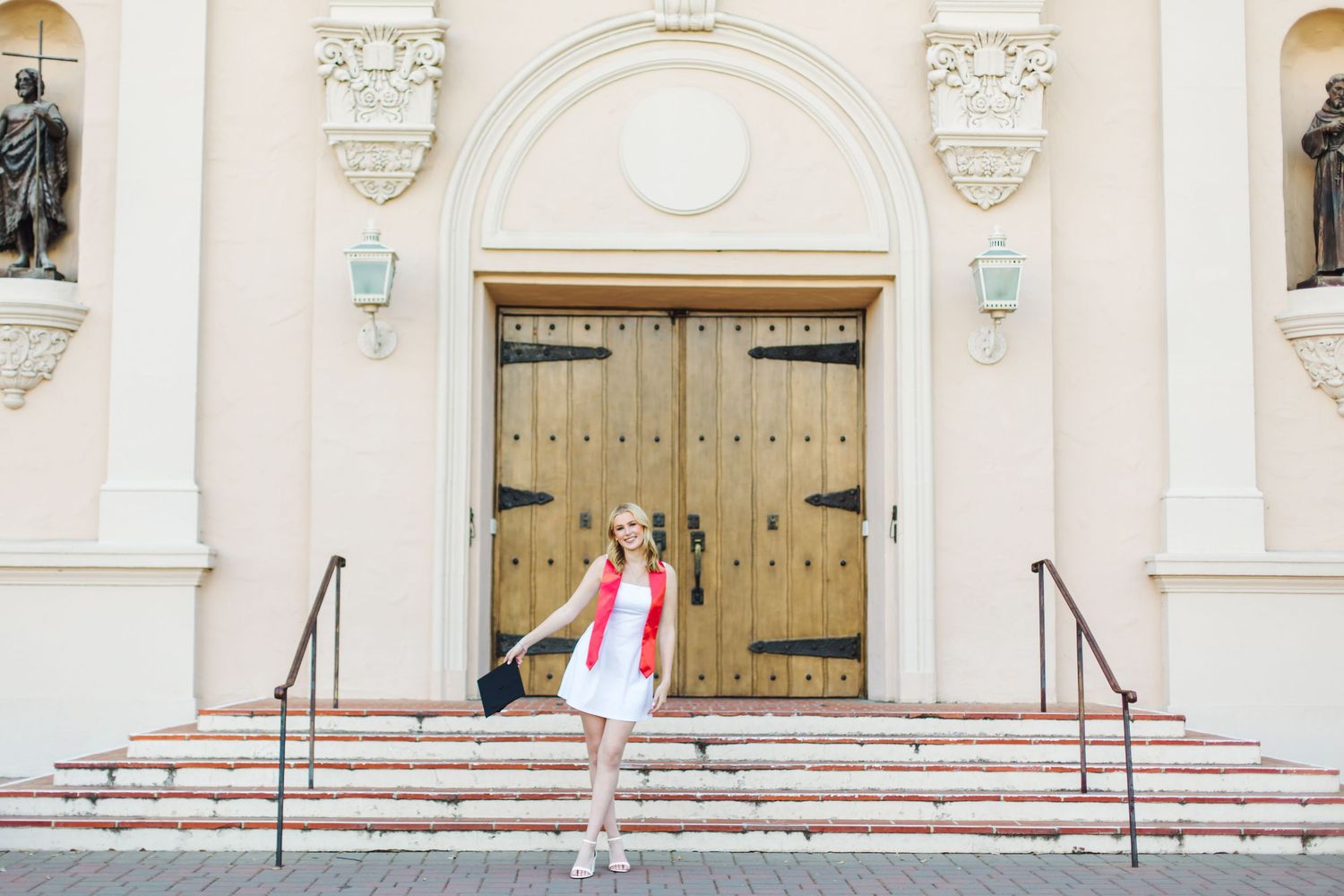 Smiling blonde graduate in white dress and red stole poses on church steps holding mortarboard.
