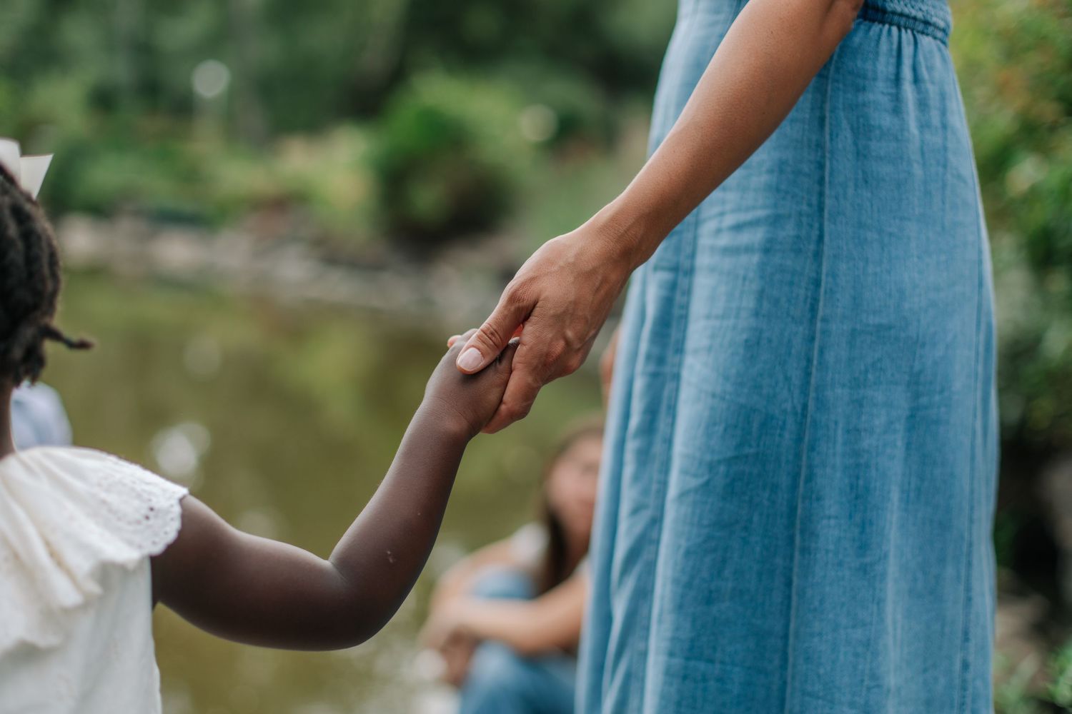 Close-up of a woman in a blue dress gently holding a young child's hand outdoors.