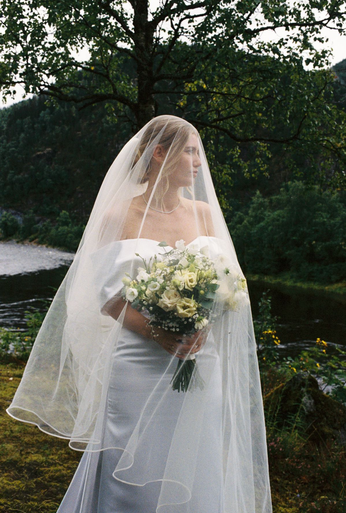 Elegant bride in a white gown and flowing veil holding a bouquet of white roses outdoors.