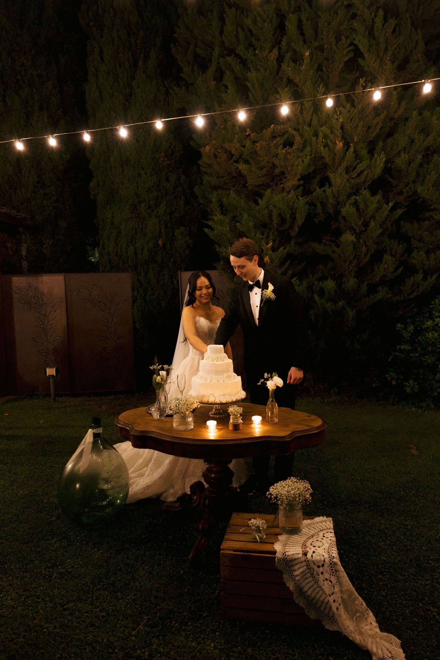 Bride and groom cutting wedding cake outdoors at night, illuminated by string lights and candles in a garden setting.