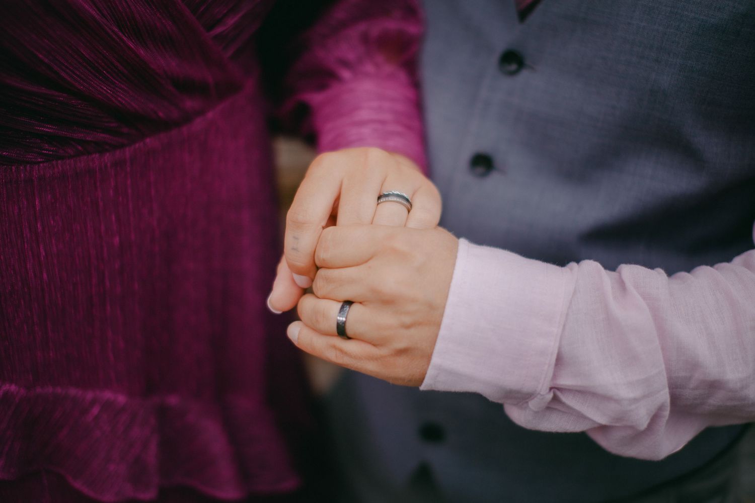Close-up of a couple holding hands, both wearing wedding bands, dressed in purple and gray formal attire.