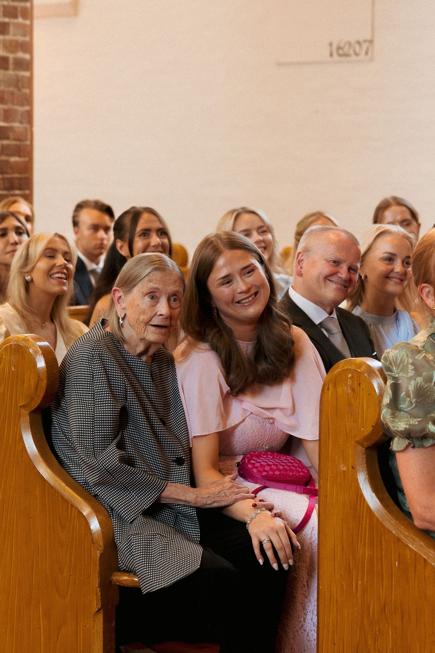 Wedding guests seated in church pews smiling, elderly woman and young woman in pink dress.