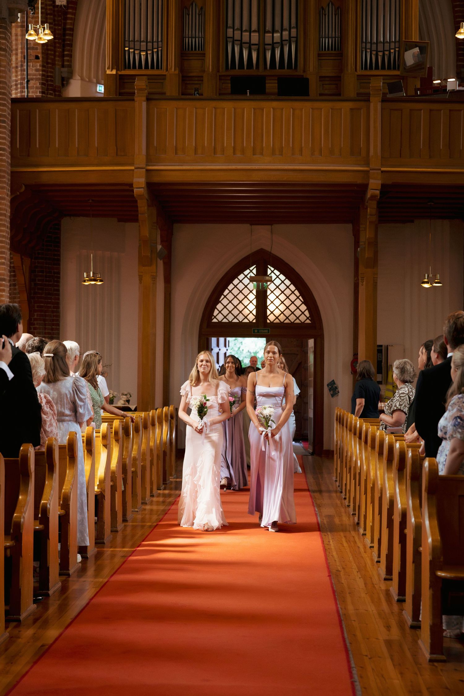 Bridesmaids walking down red carpet aisle in wooden church during wedding ceremony.