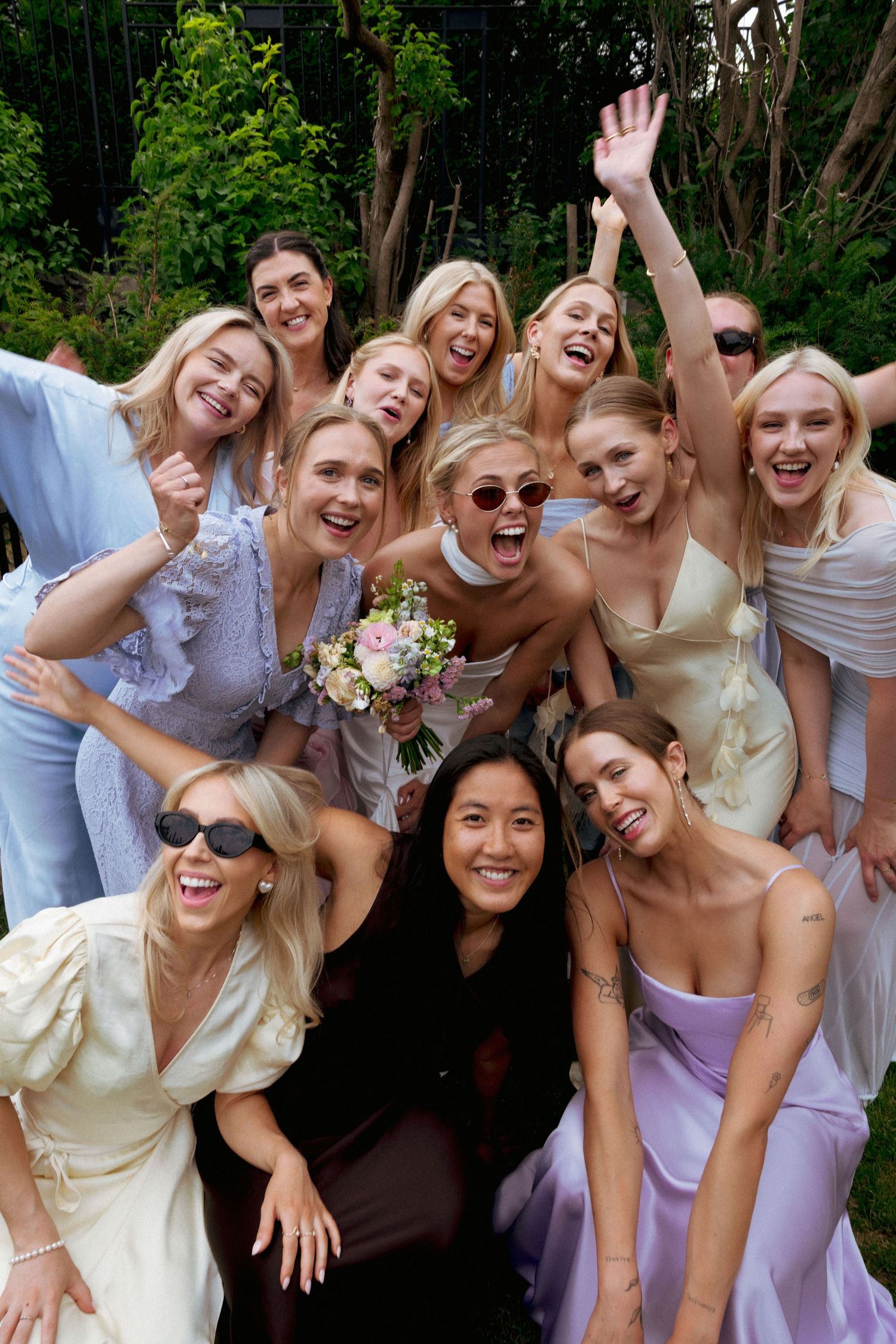 Bride holding bouquet celebrates with bridesmaids in pastel dresses outdoors.